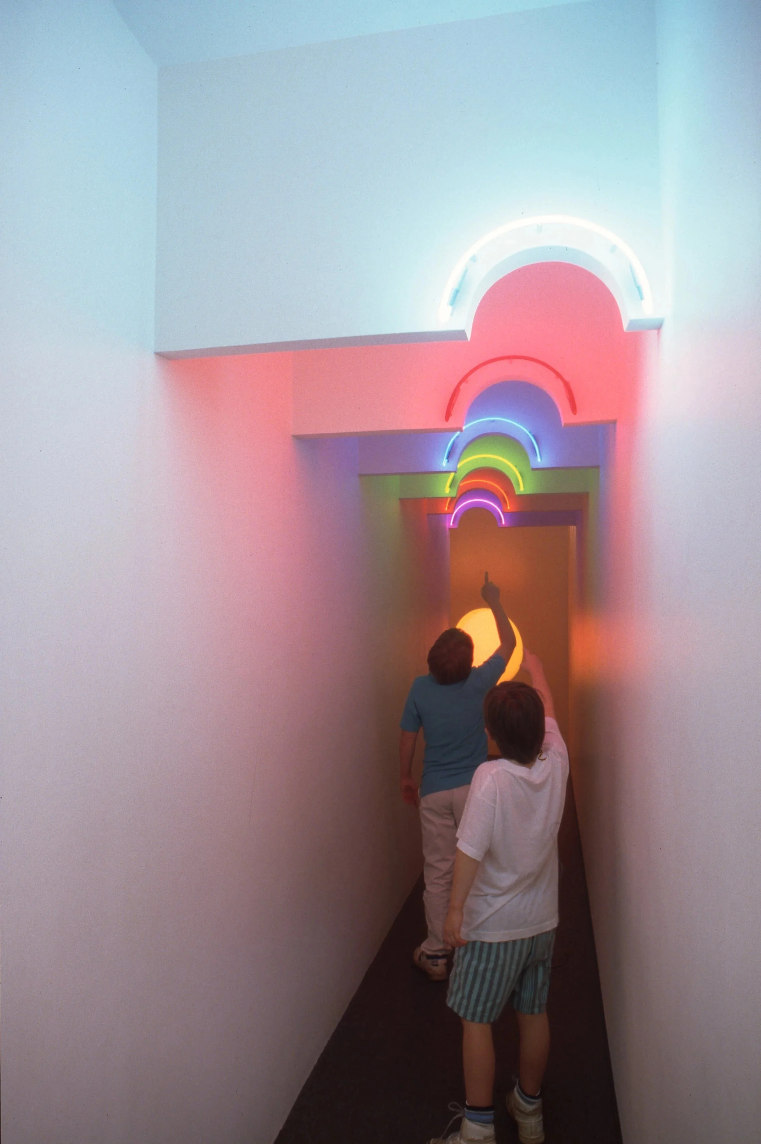 Two children walking through a hallway with colorful neon rainbow lights on the ceiling.