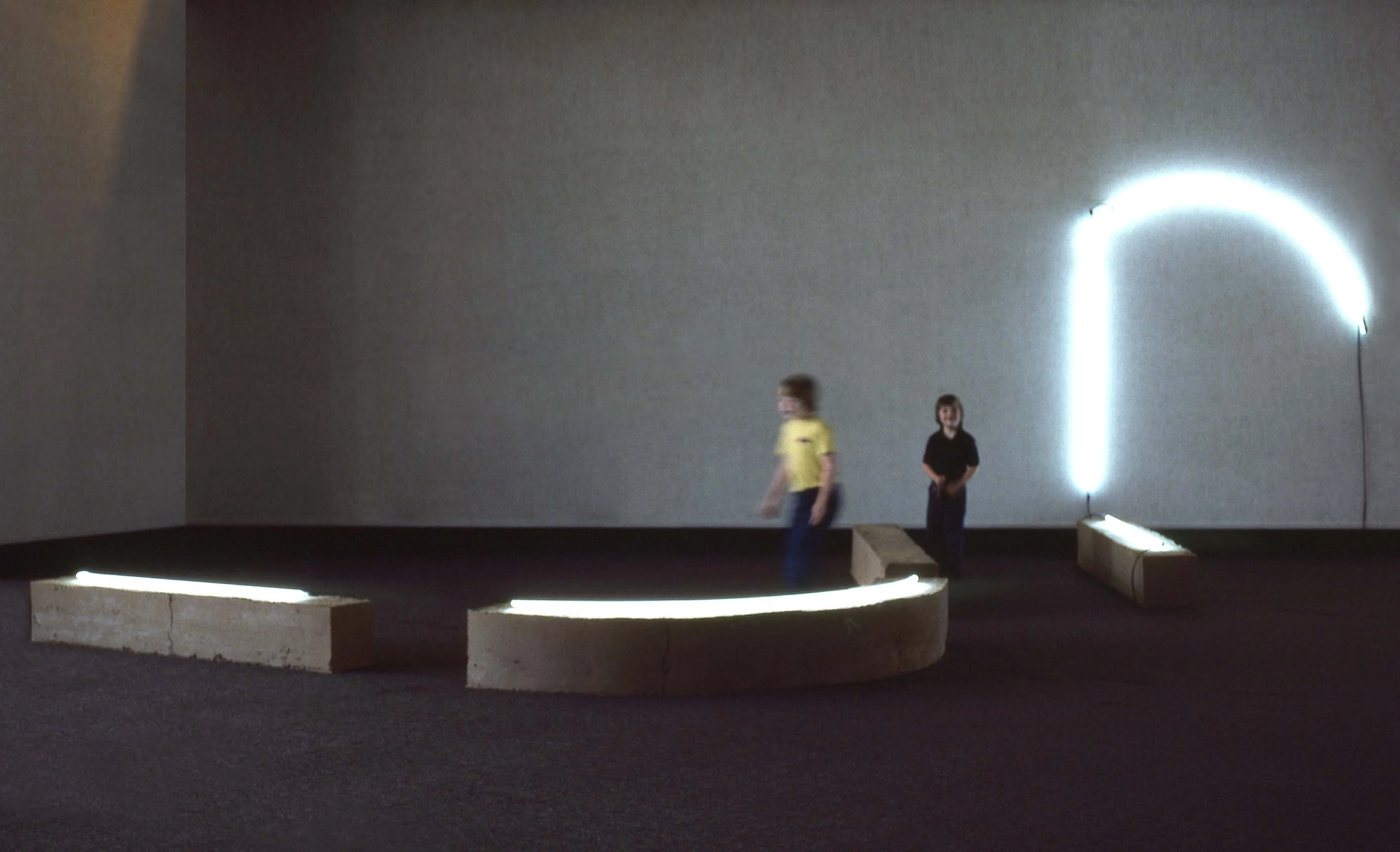 Art installation with curved concrete benches outlined with white LED lights and a neon light arch or question mark on a plain wall, with two children interacting.