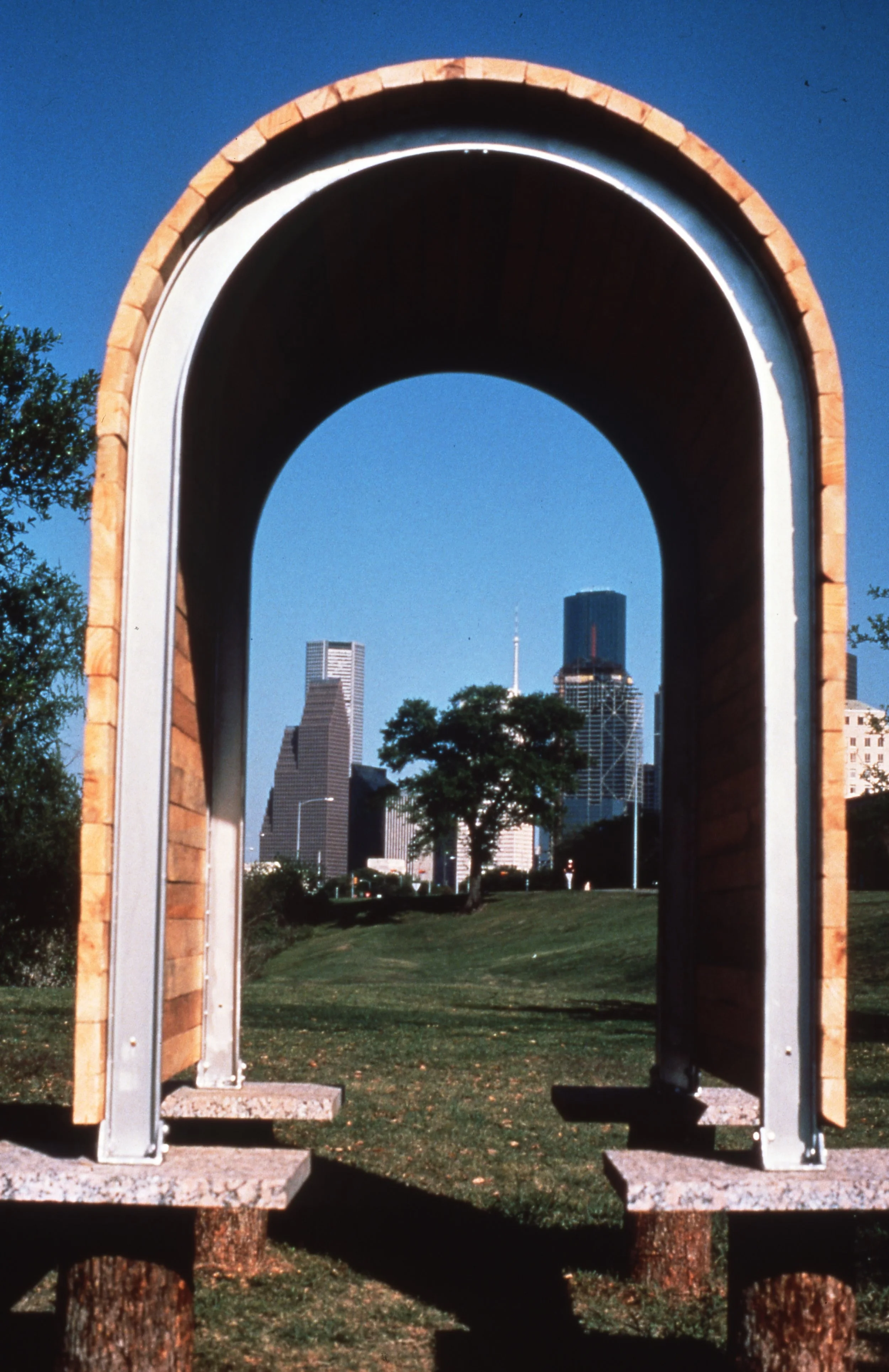 A park bench shaped like an open book with a city skyline in the background, seen through a large stone and metal archway.