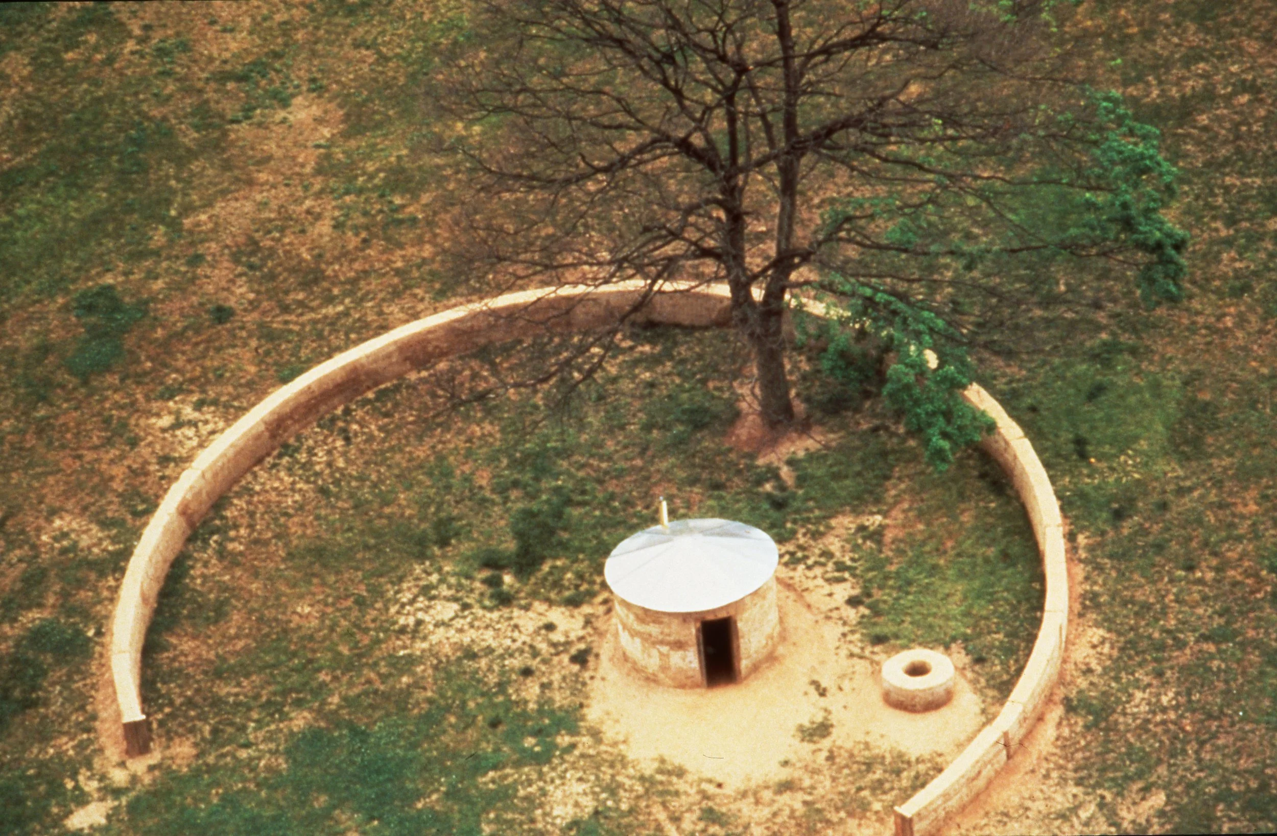 An aerial view of a small circular stone structure with a white roof, surrounded by a curved stone wall and a tree, in a grassy area.