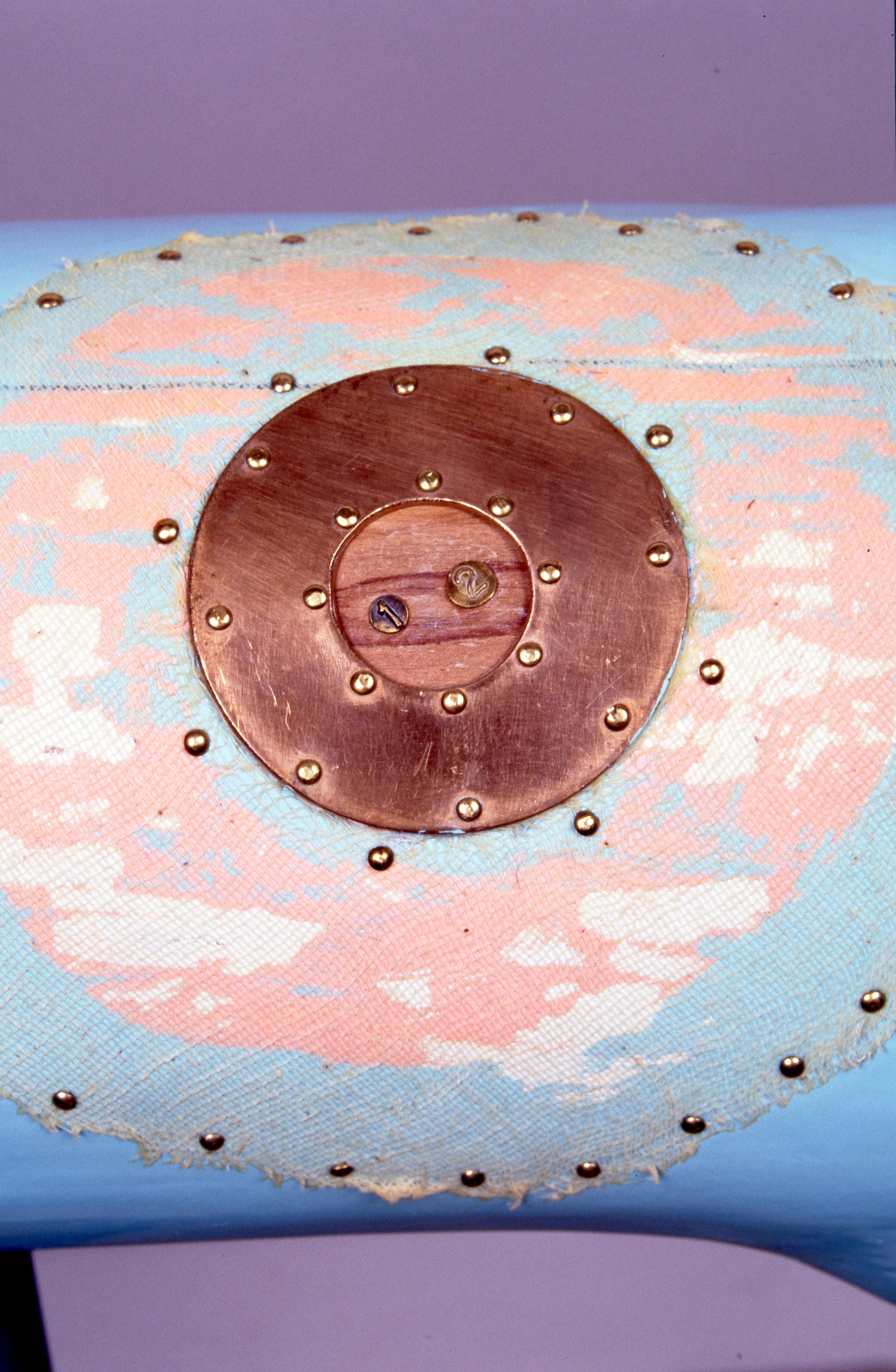 Close-up of a decorative item with a lace fabric featuring a floral pattern, topped with a round metallic disc and small beads, and embossed with numbers '1' and '2'.