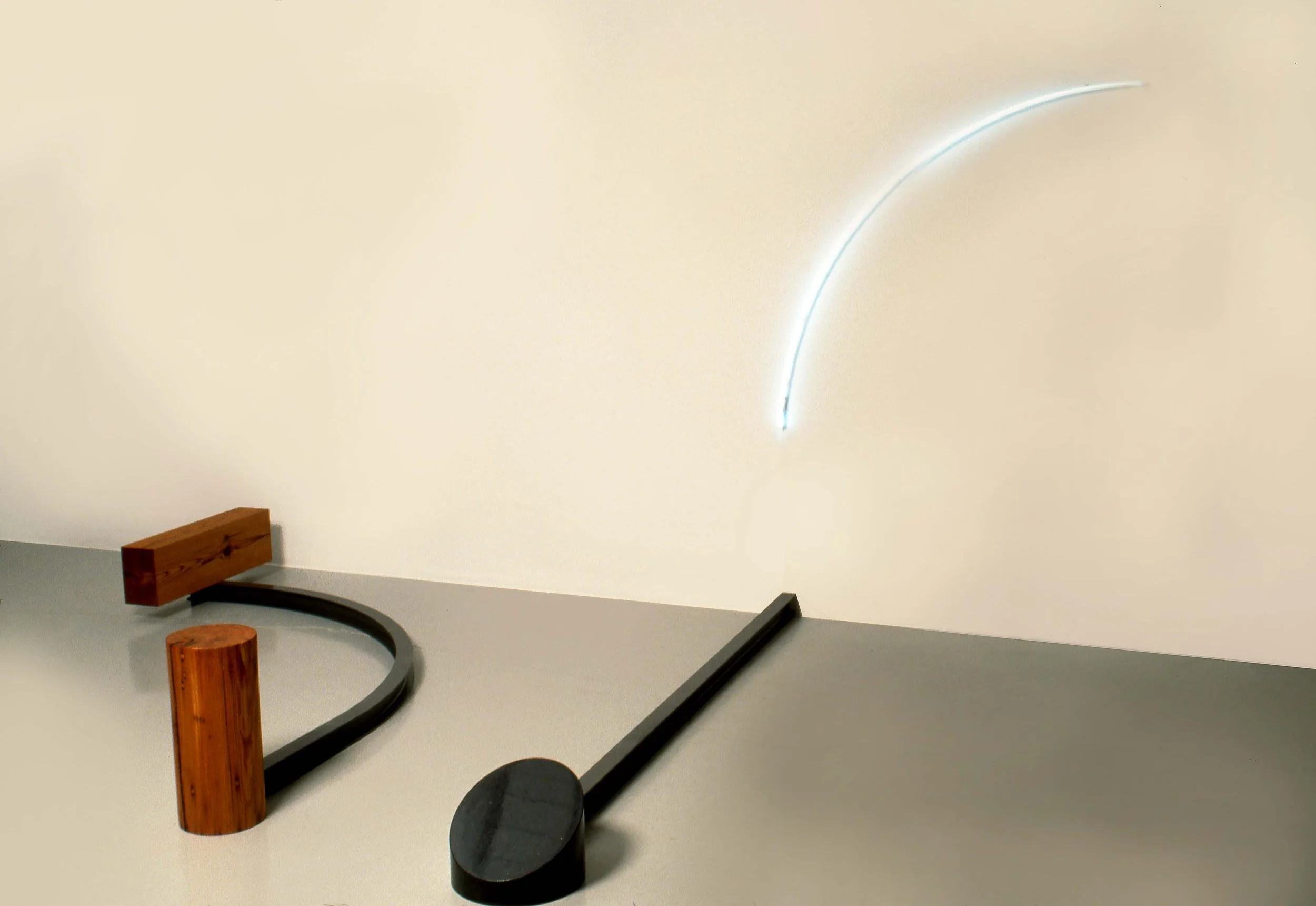 Abstract mixed media art installation with wooden and metal objects and a glowing arc on a white wall.