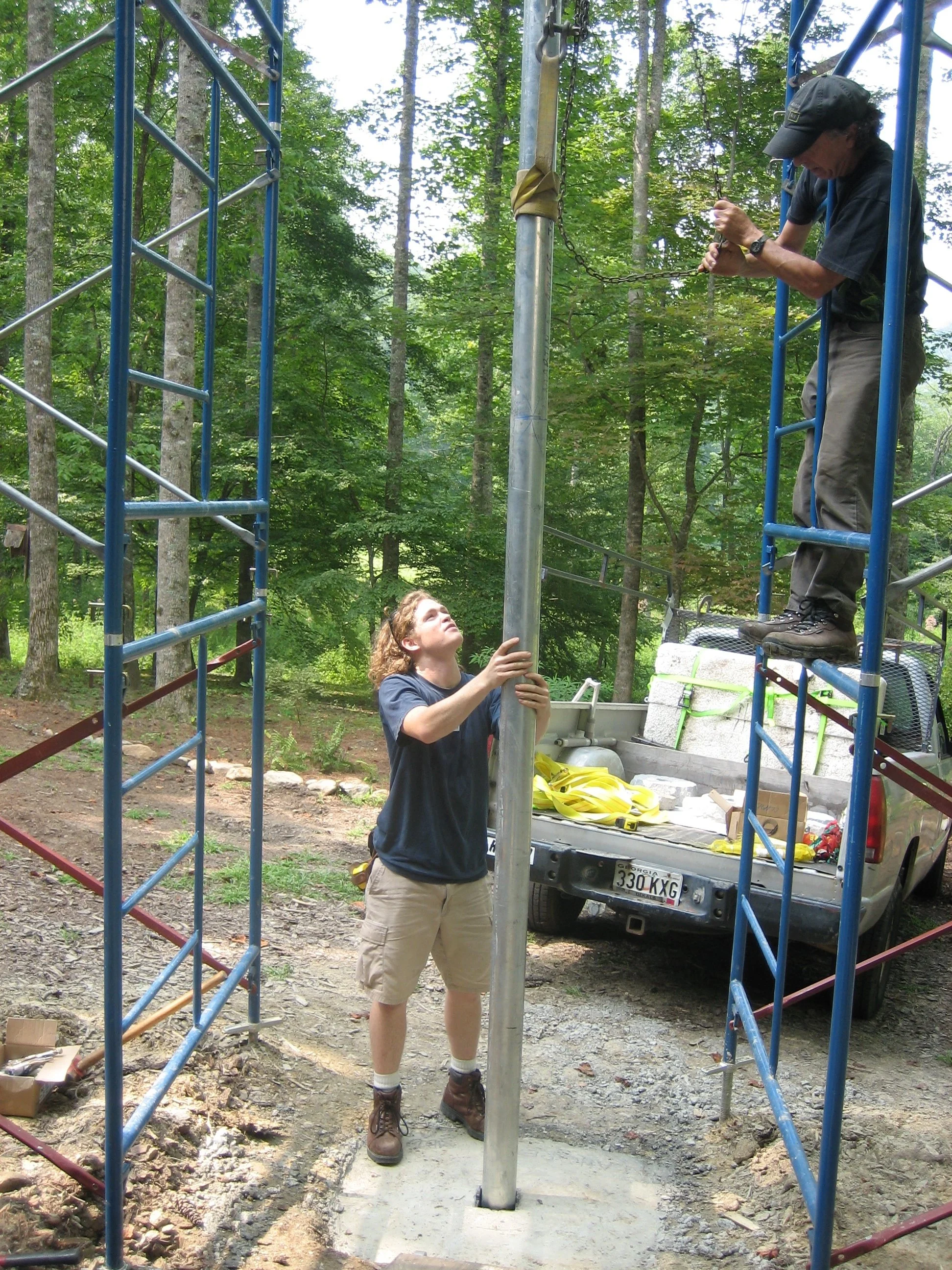 Two people assembling a metal structure outdoors in a wooded area, with a truck nearby. One person is standing on the ground holding a pole, while the other is standing on the structure securing a chain to a vertical pole.