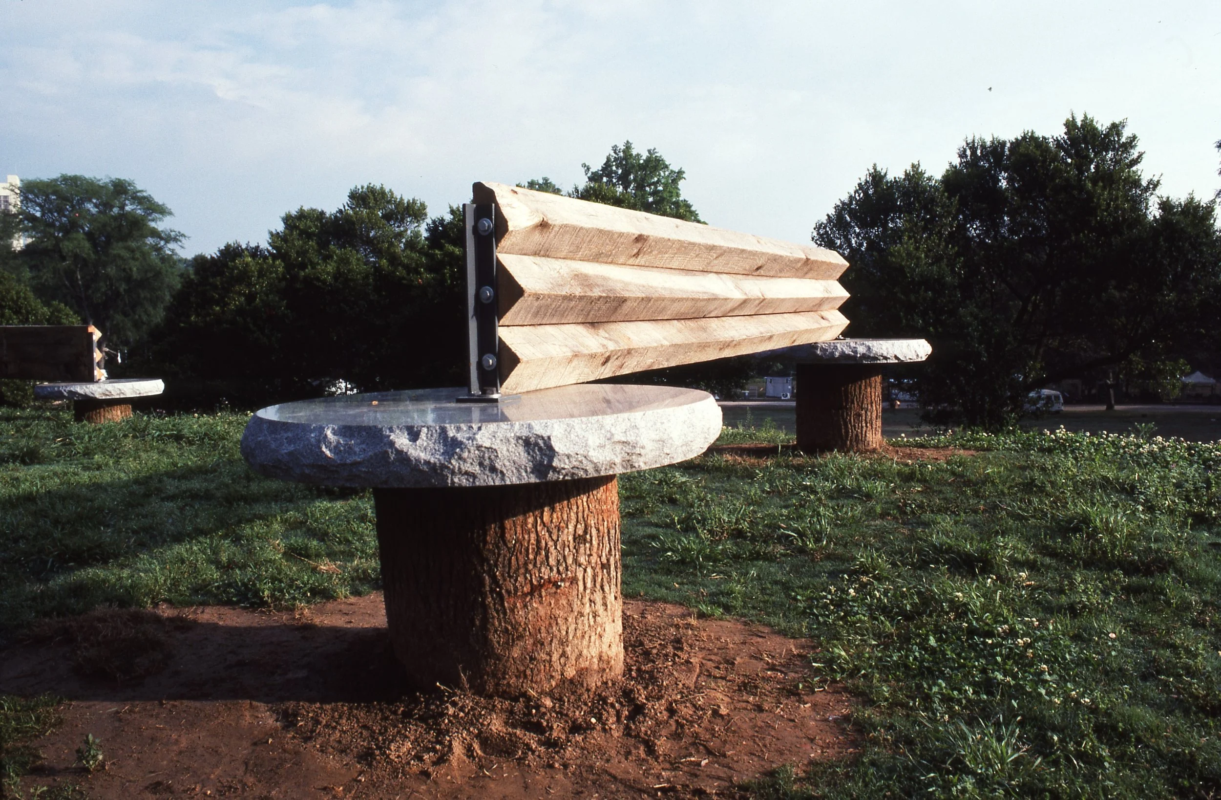 An outdoor park sculpture of a large saw made from wood slats and metal bolts, mounted on a stone and log base, with green trees and a clear sky in the background.
