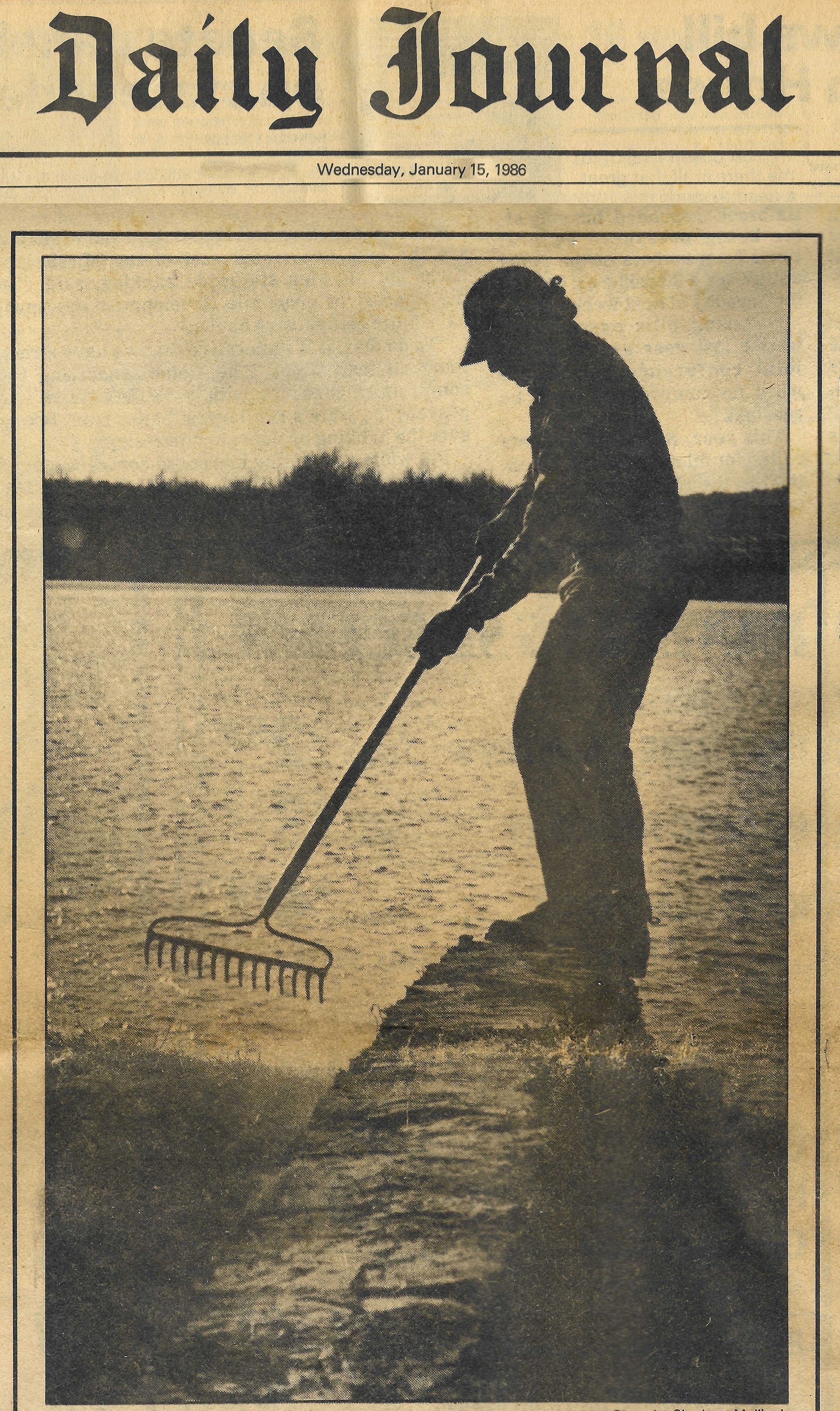 A silhouette of a person wearing a hat, standing on a log by a body of water, raking the water's surface, during sunset or sunrise.
