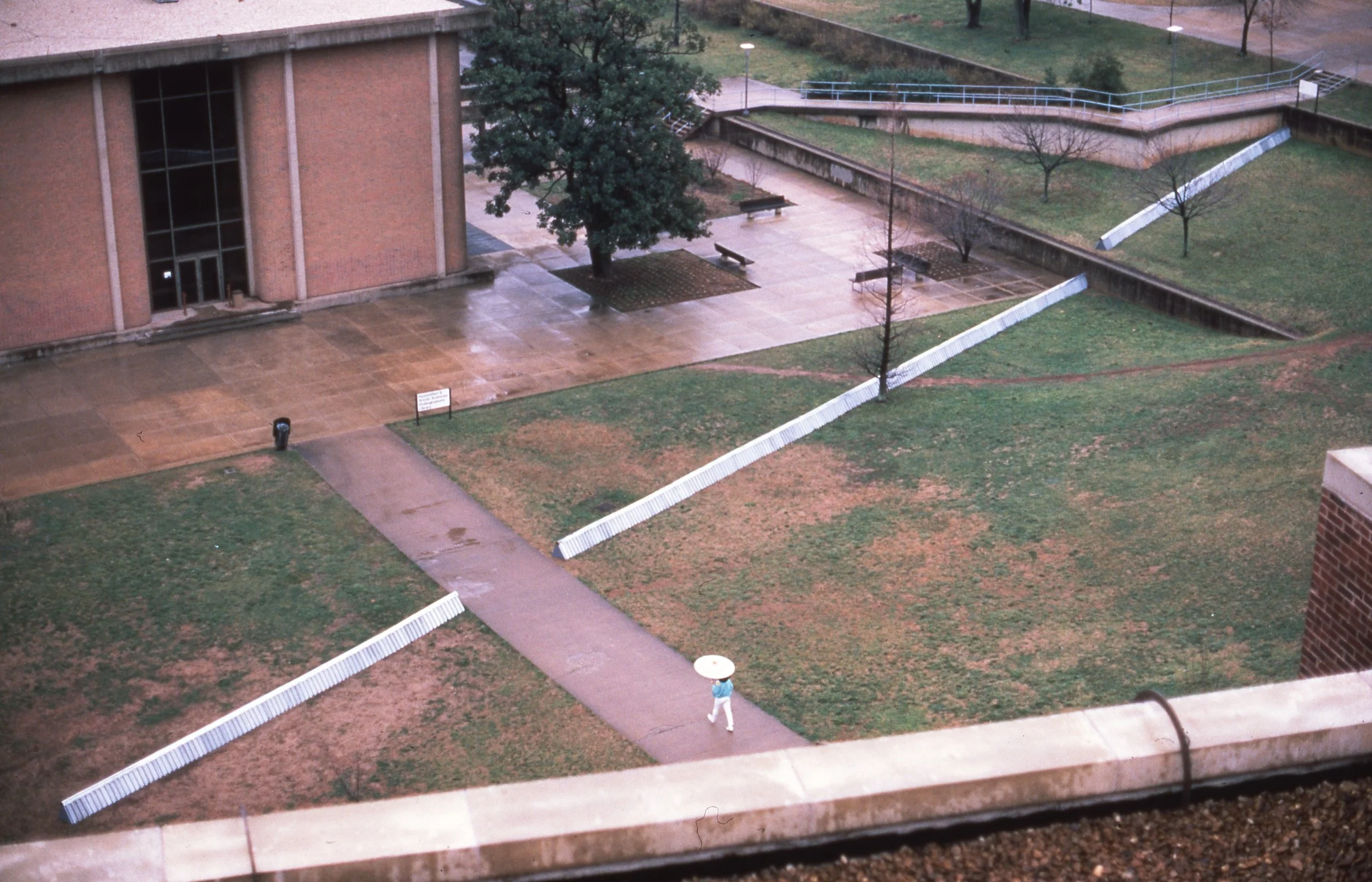 Empty campus yard with benches, trees, a person walking with an umbrella, and a building.