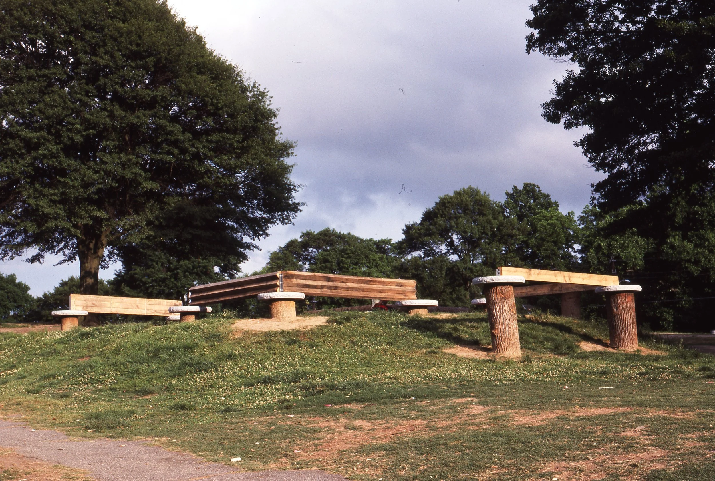 Empty public park with wooden benches on a grassy hill, surrounded by large green trees under a cloudy sky.