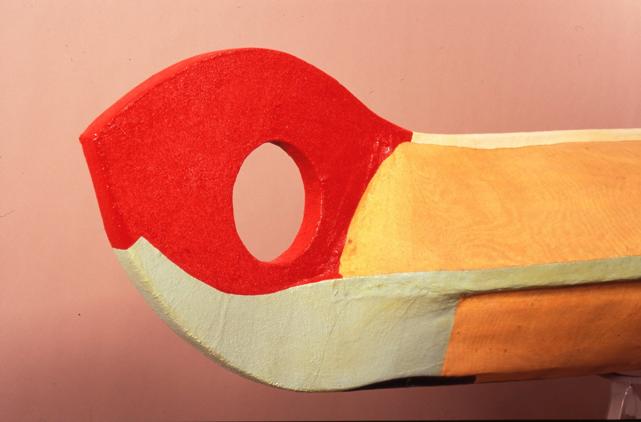 Close-up of a colorful wooden toy resembling a cartoonish drill with a circular hole at the tip, painted in red, yellow, and white, against a pink background.