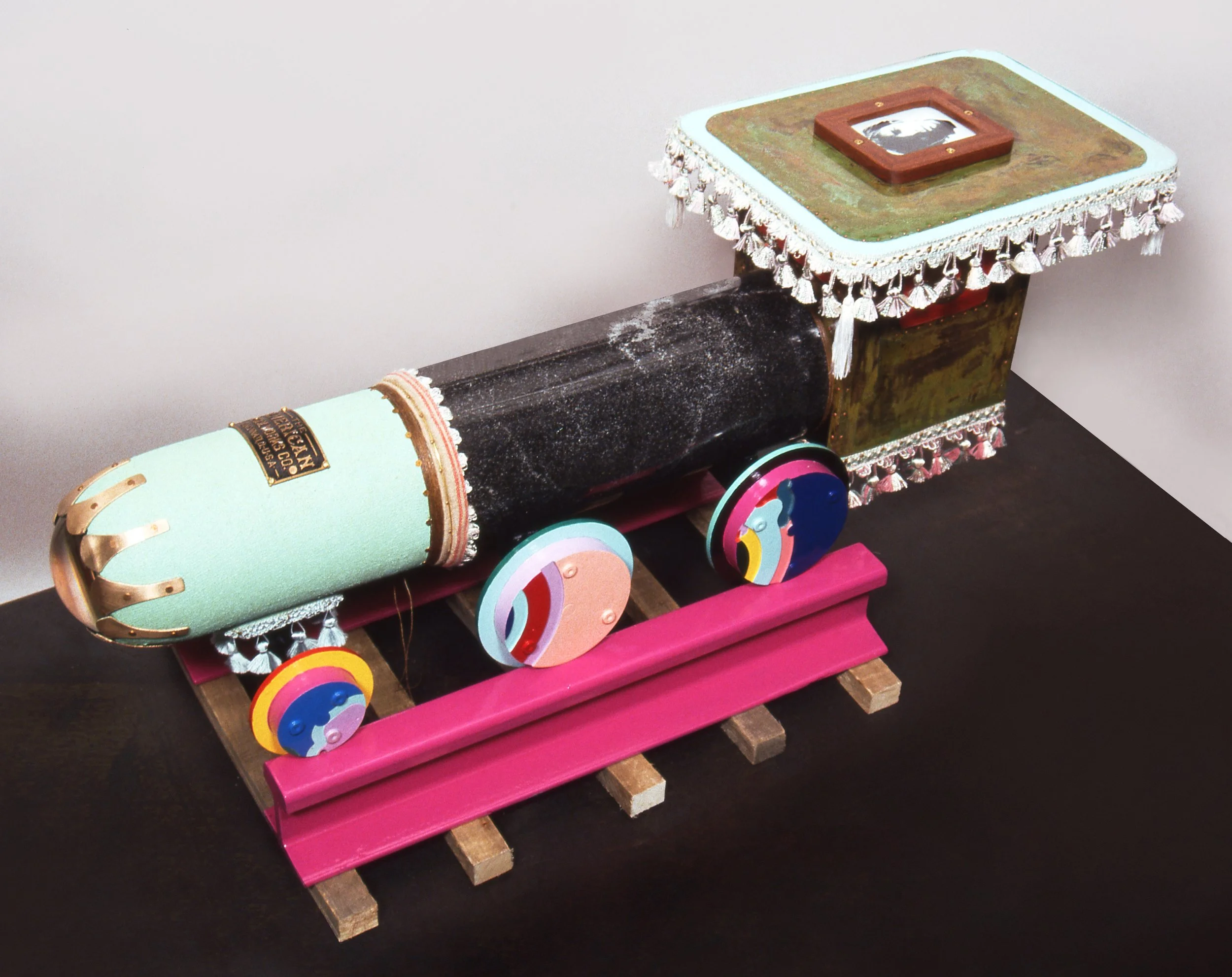 Colorful model of a train on a pink wooden base with decorative wheels, featuring a black and green body with ornate trim, and a green and brown top with decorative tassels.