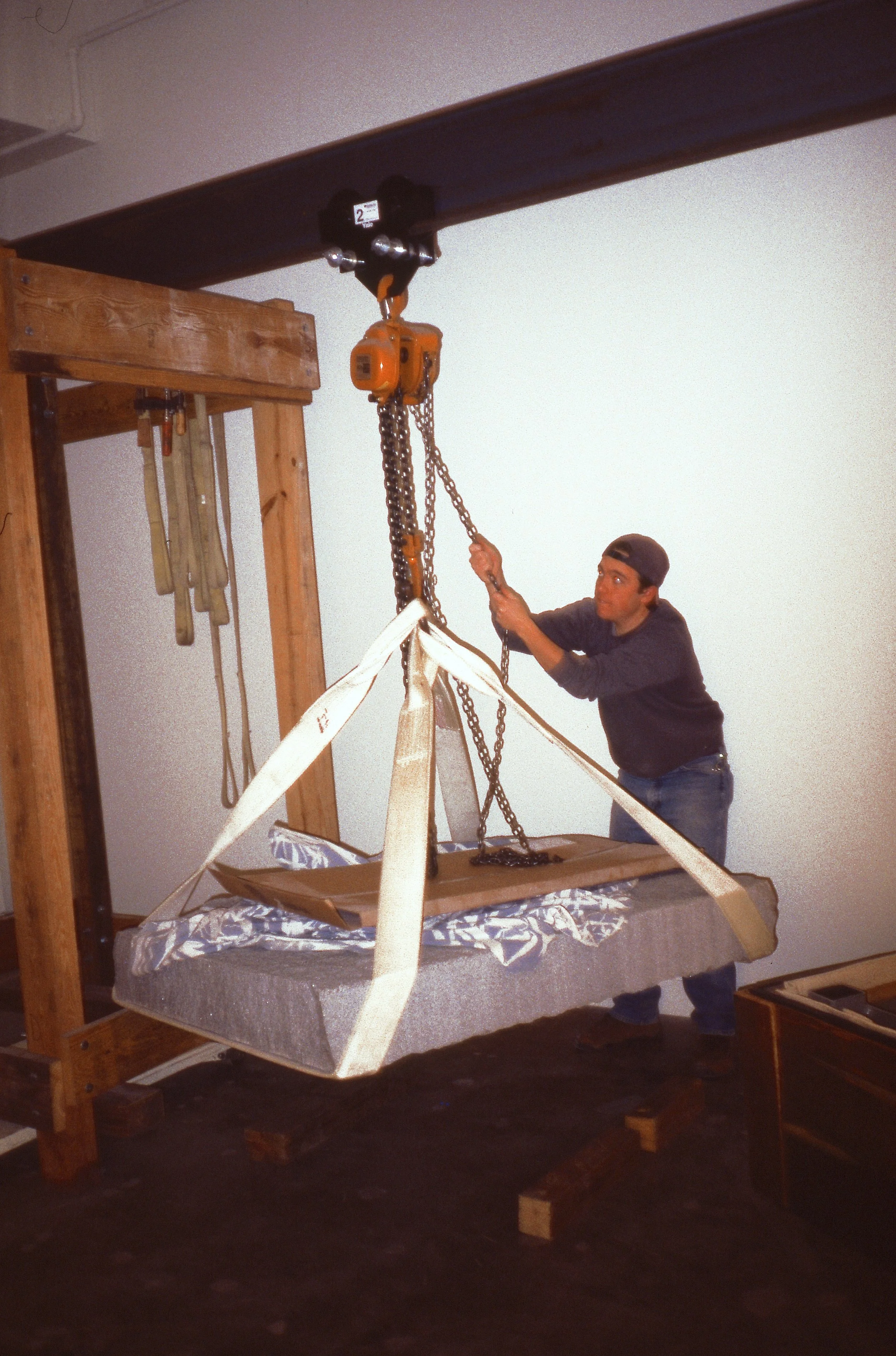 Man operating a hoist to lift a large, wrapped piece of furniture in a workshop.