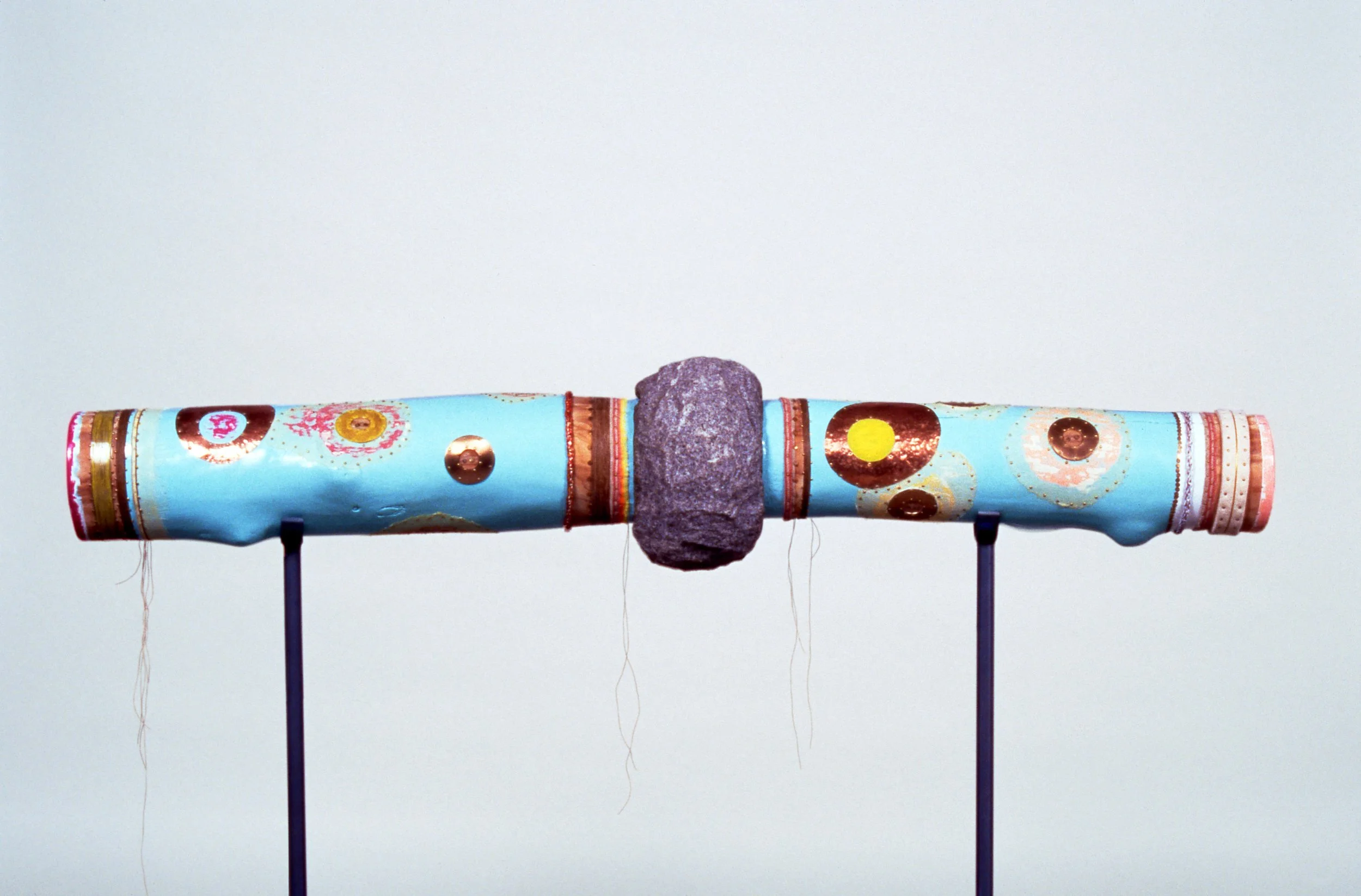 Mixed-media artwork on a horizontal cylindrical surface, supported by two black stands, featuring a central stone, colorful patterns, circular designs, and metallic accents on a light blue background.