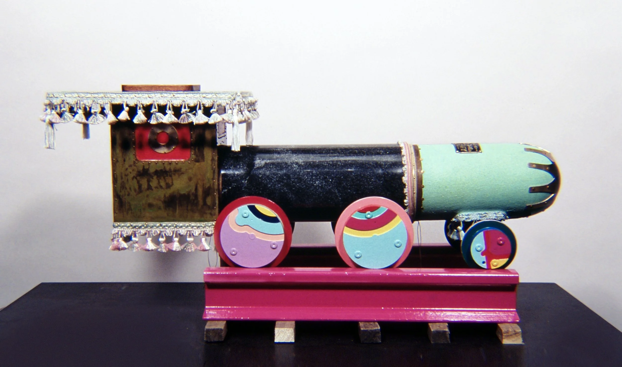 A miniature train model with decorative elements, colorful wheels, and a combination of black, pink, and mint green colors, placed on a wooden surface.