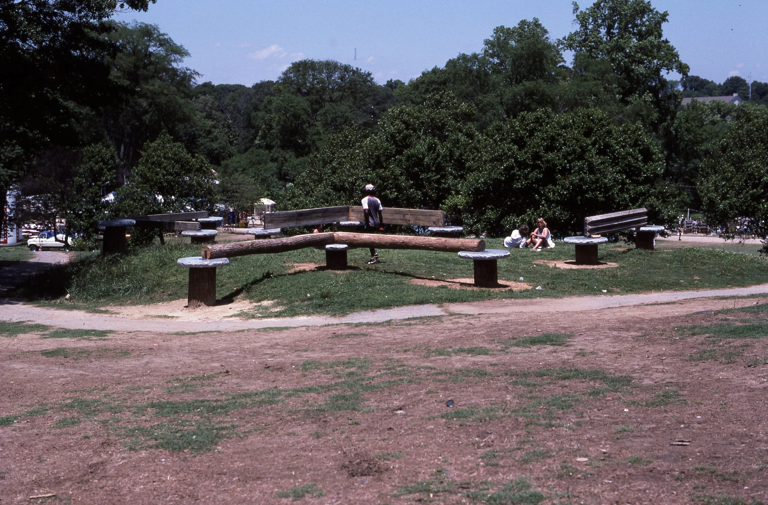 A park with trees and open grassy areas, a balance beam made from logs, and a few children playing or sitting on the grass.