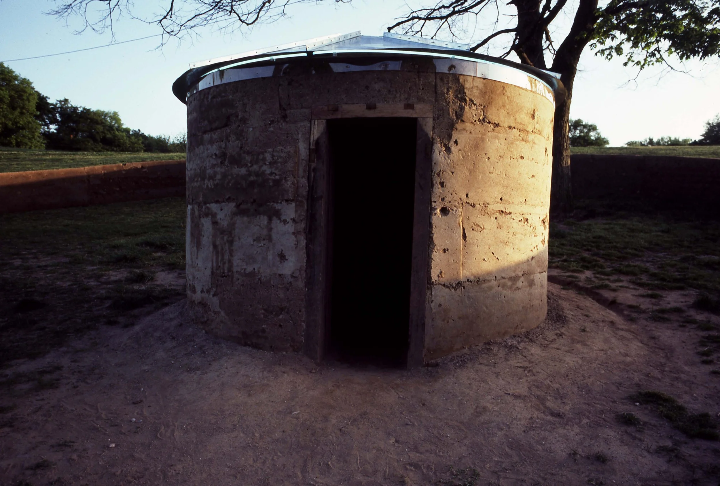 A round, stone structure with an open doorway, outdoors with trees and grass in the background, under clear sky.