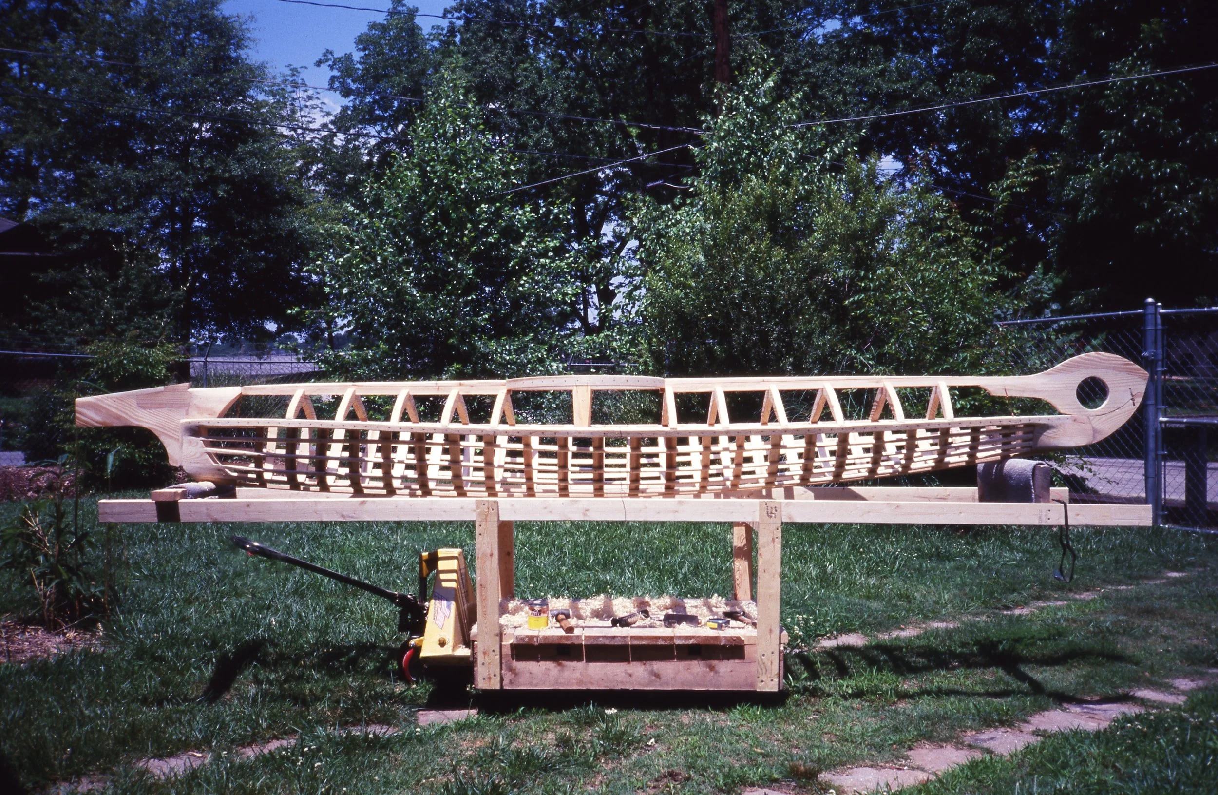 Wooden boat model under construction on a workbench outdoors, with trees and a fence in the background.