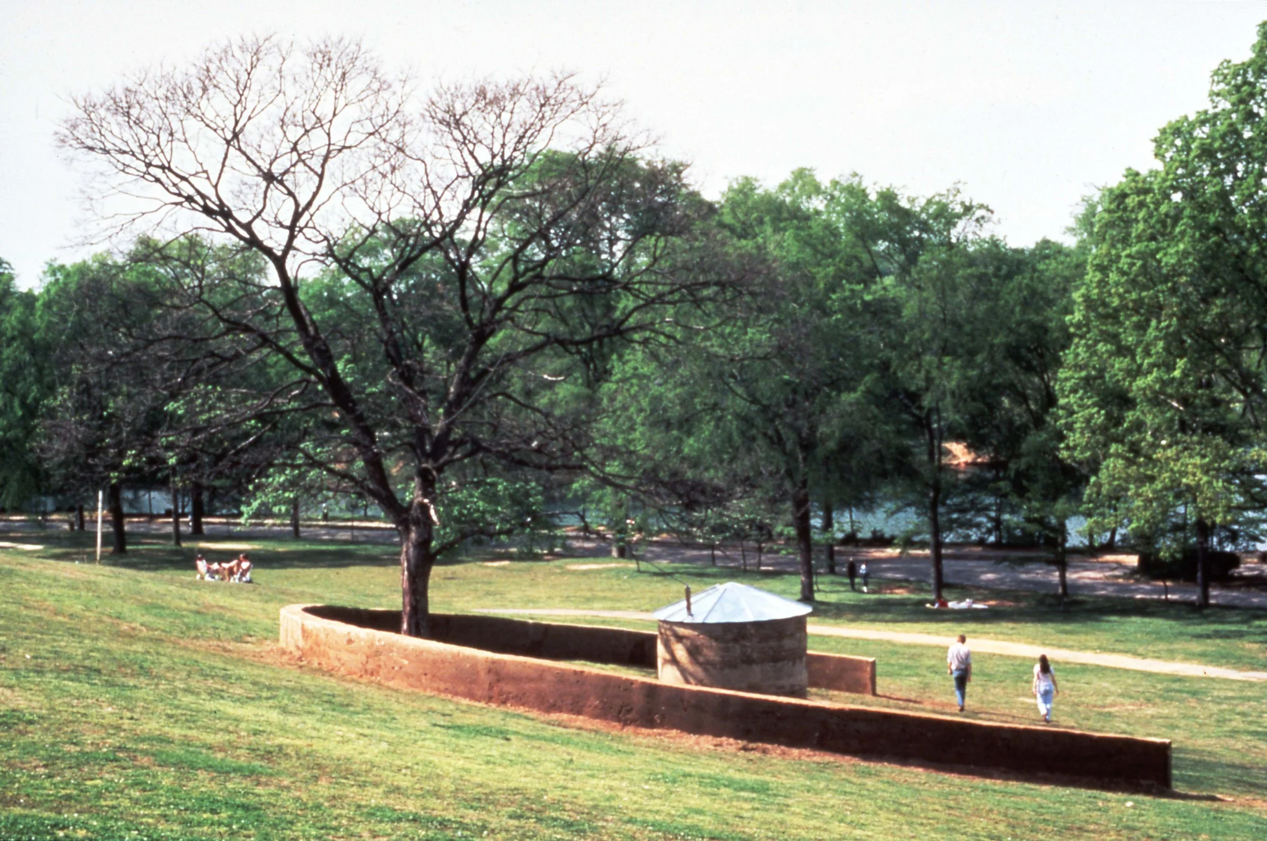 A park scene with a large leafless tree, green trees, a stone well with a metal roof, and people walking on a dirt path.