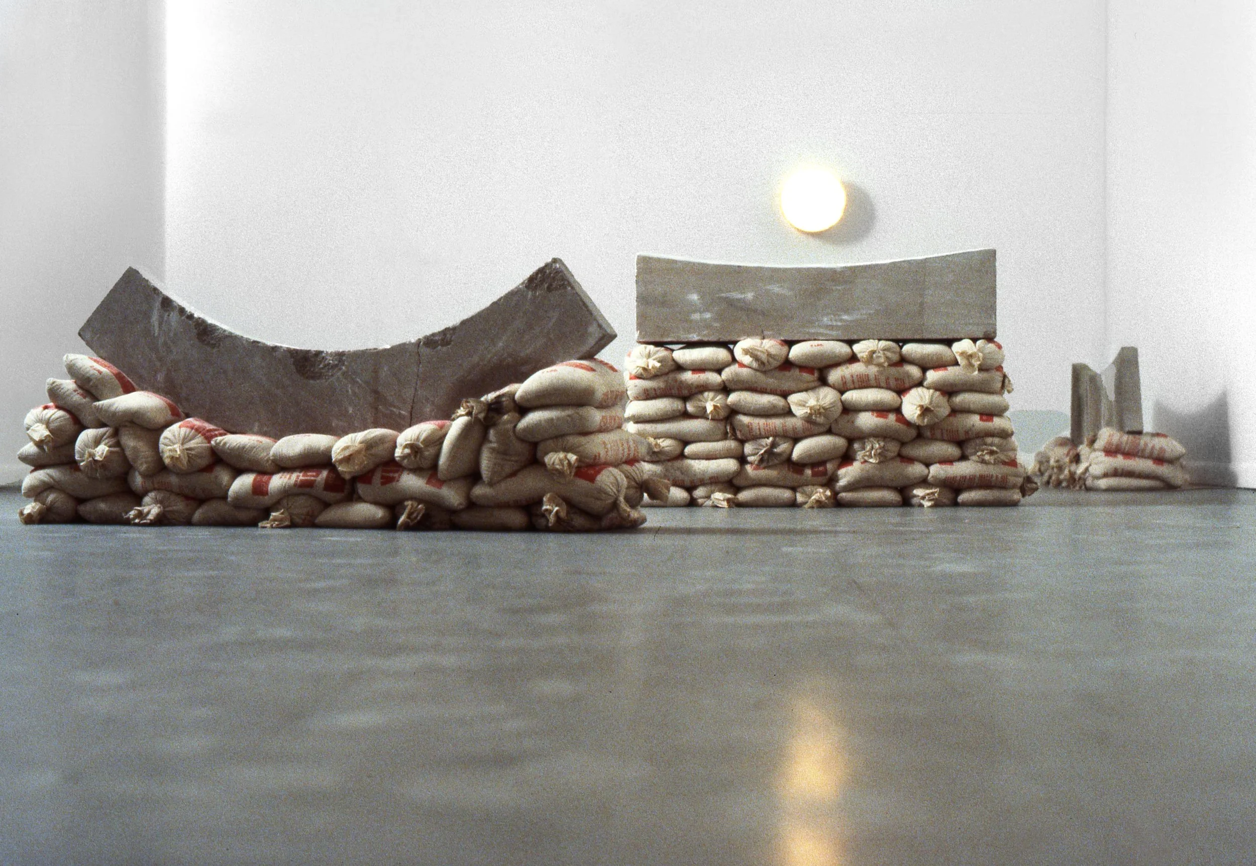 A minimalist sculpture resembling a landscape, made of stacked sandbags, a curved concrete piece, and rectangular blocks, set against a plain gray background with a suspended bright light or spotlight.