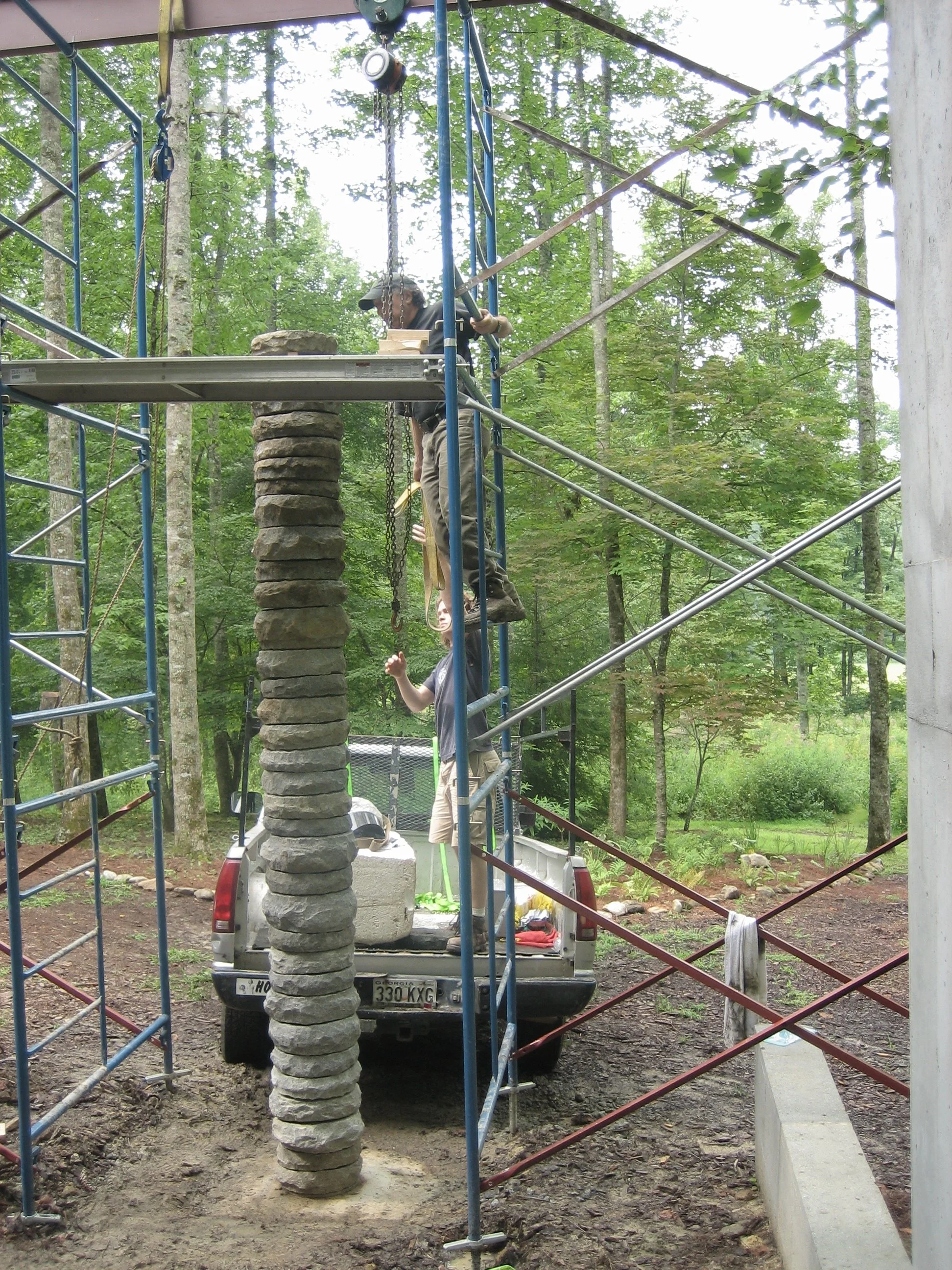 People working on outdoor construction project with scaffolding, truck, and stone pillar in a wooded area.