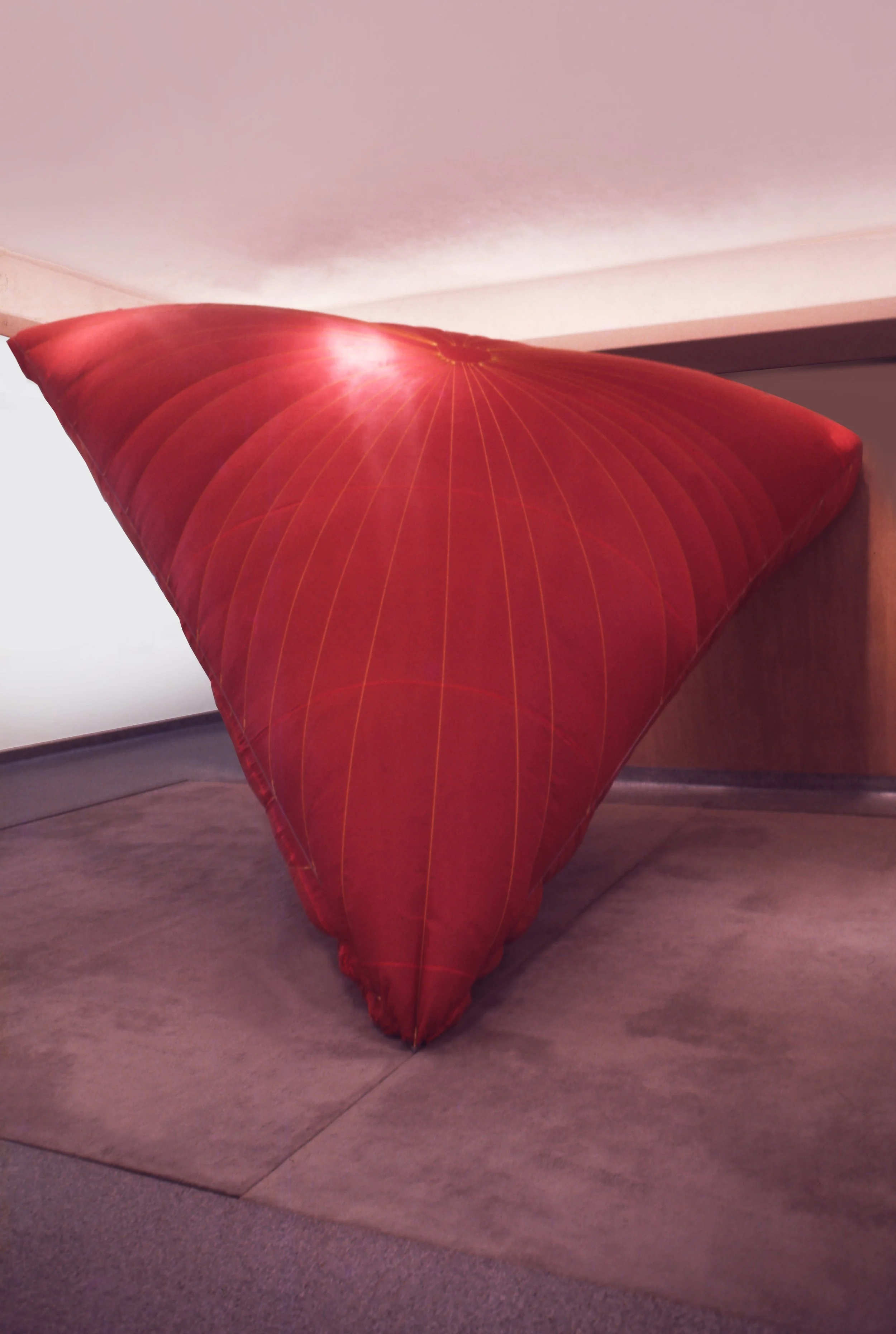 A large, red, folded fabric structure resembling a tent or canopy positioned on a wooden floor in a room with pink walls.