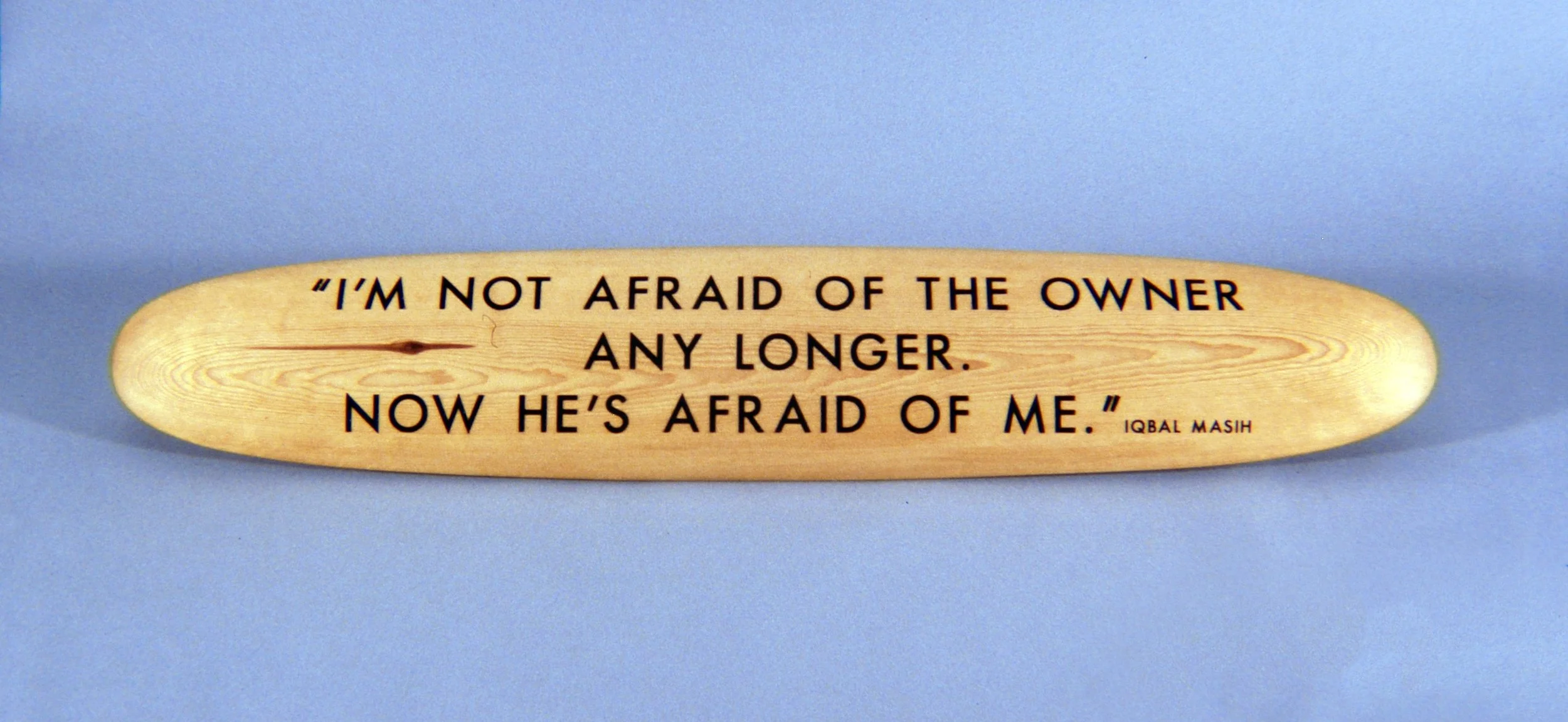 A wooden paddle with a quote by Iqbal Masih painted on it, reading: "I'm not afraid of the owner any longer. Now he's afraid of me."