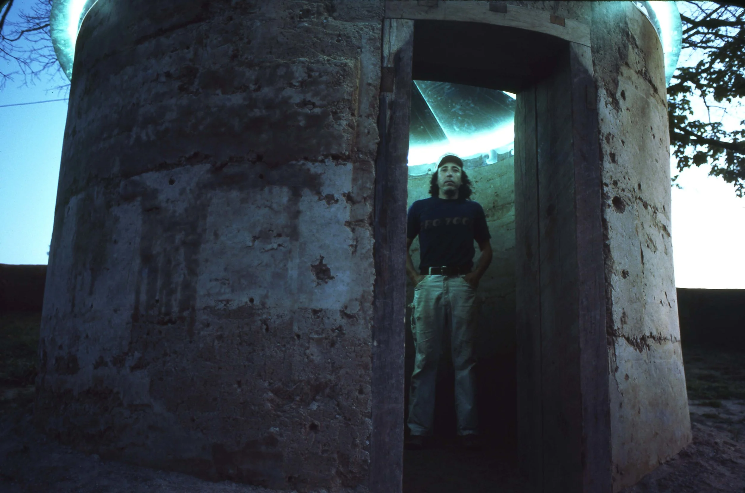 A man standing inside a small, cylindrical stone structure with an open door, illuminated by greenish-blue lighting, with trees visible in the background.