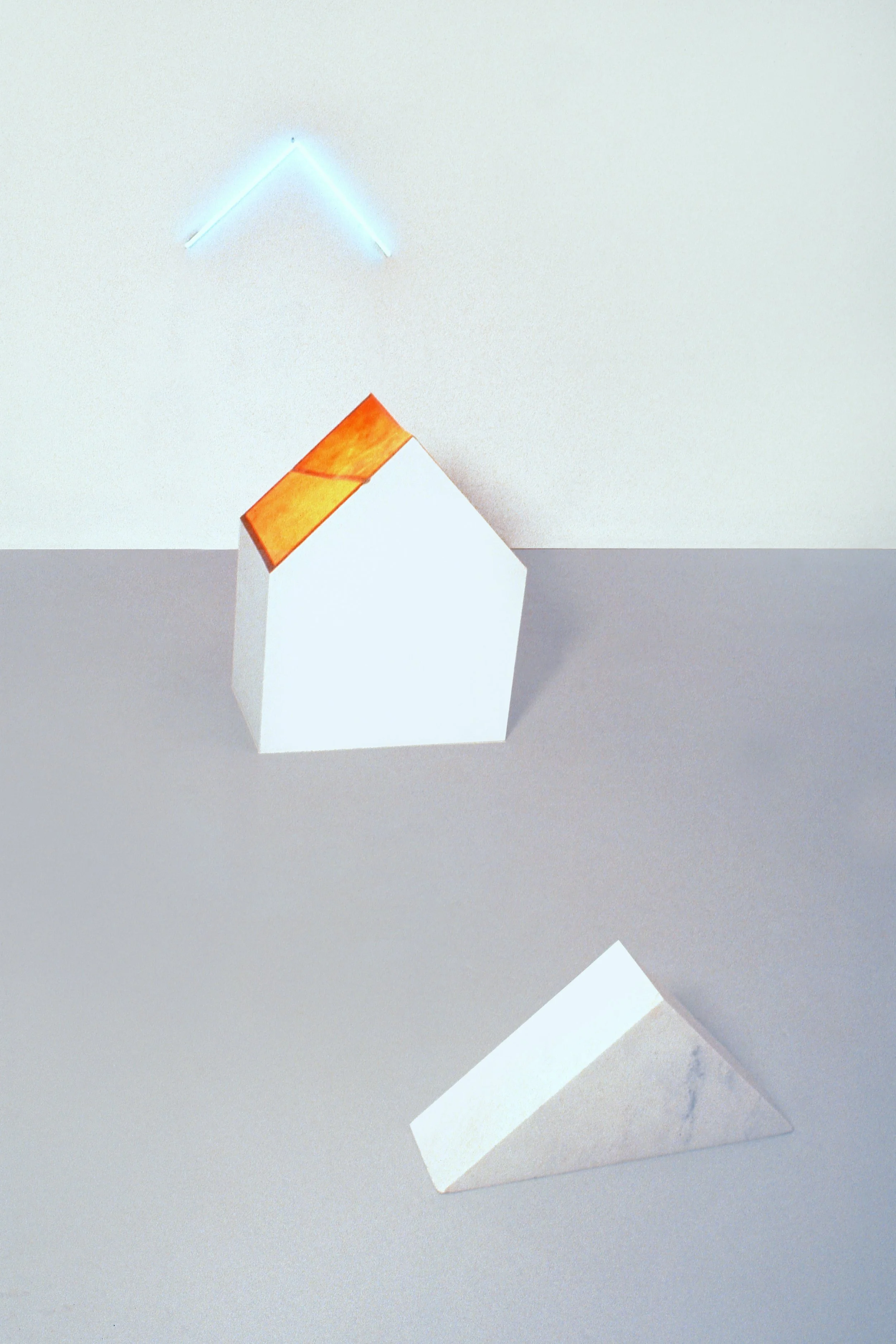 A minimalist composition with a white geometric house shape with an orange roof, a white marble rectangular block leaning against a light grey surface in front of a white wall, and a glowing blue triangular light above.
