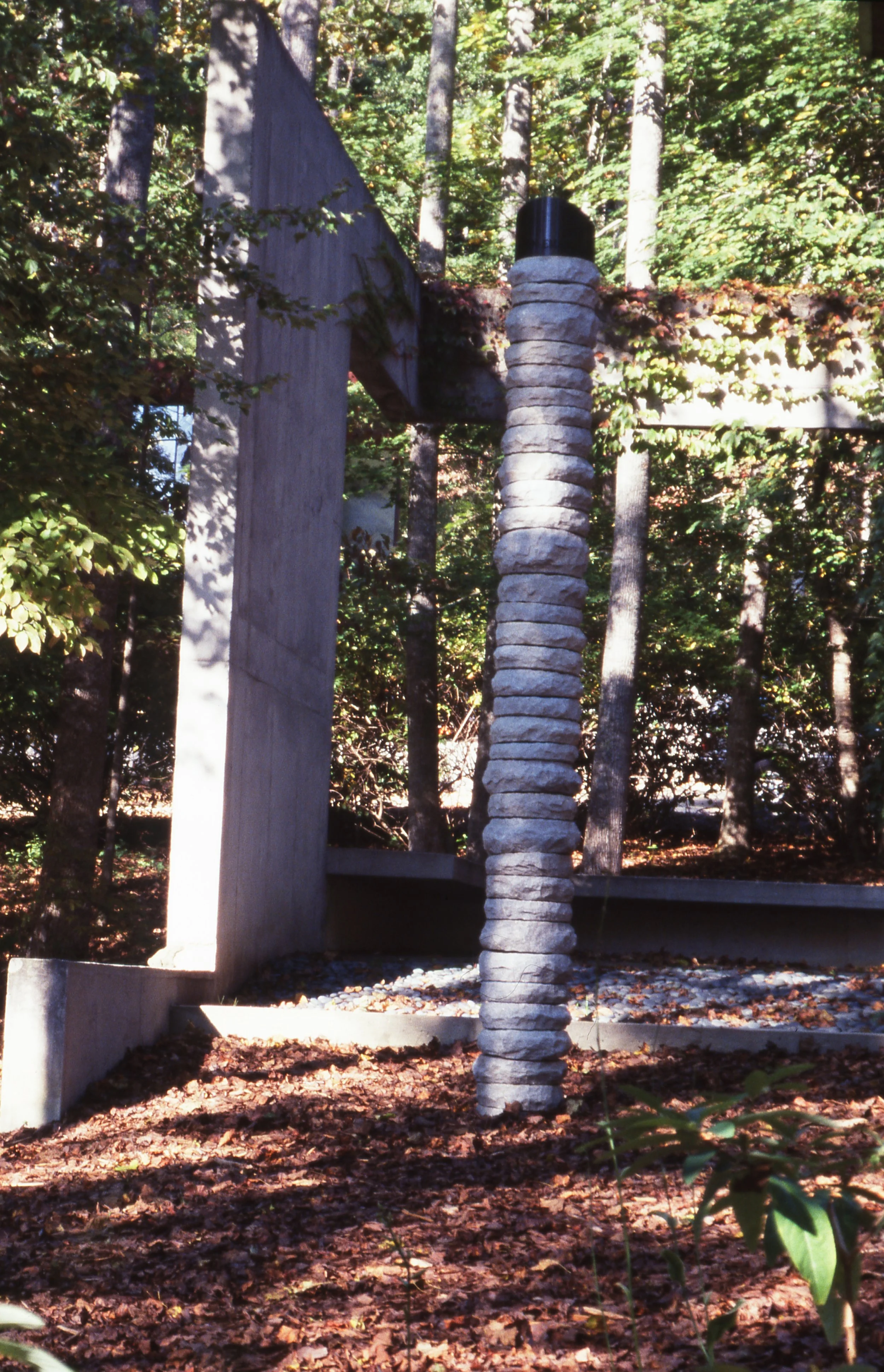 Outdoor sculpture composed of stacked flat stones on a wooden post, surrounded by trees with green leaves and fallen autumn leaves on the ground.