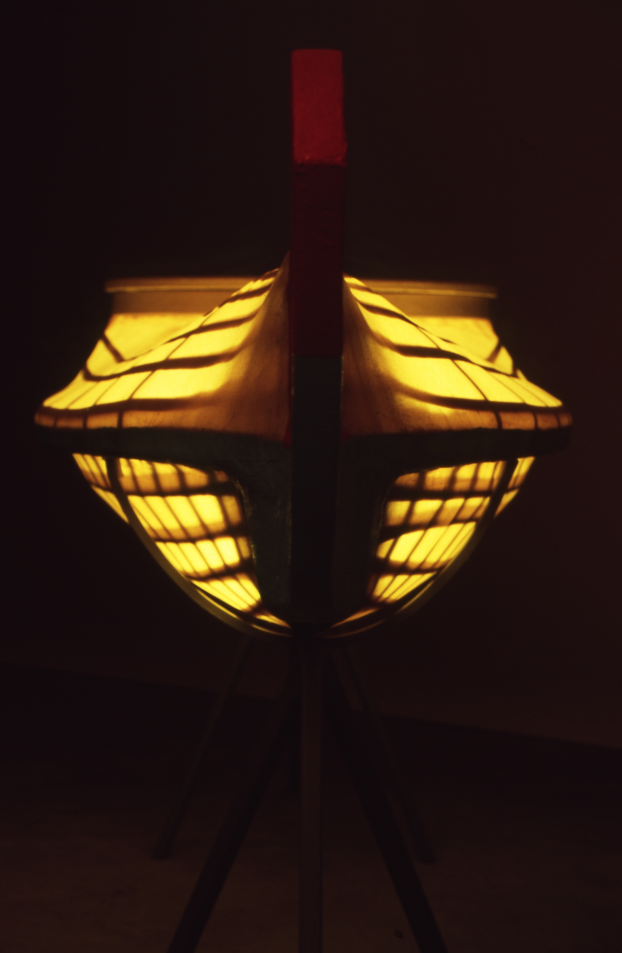 A ship-shaped yellow lampshade with black trim, illuminated from within, standing on three legs in a dark room.