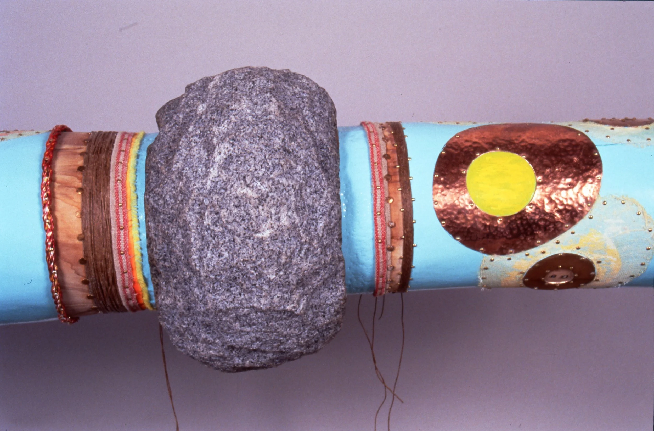Decorated cylindrical object with colorful designs, a large gray rock attached to it, and a prominent copper-colored circle with a yellow center, set against a plain background.