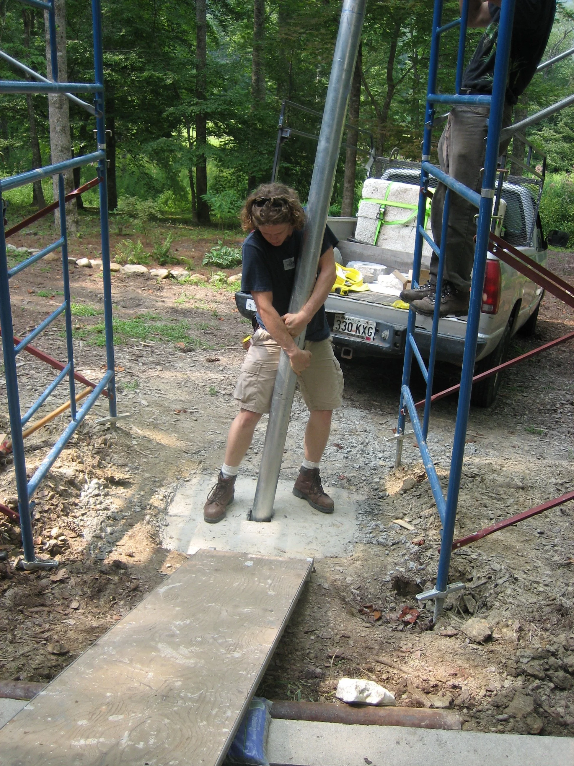 Person working on installing a metal pole into a concrete base outdoors with scaffolding and a truck in background.