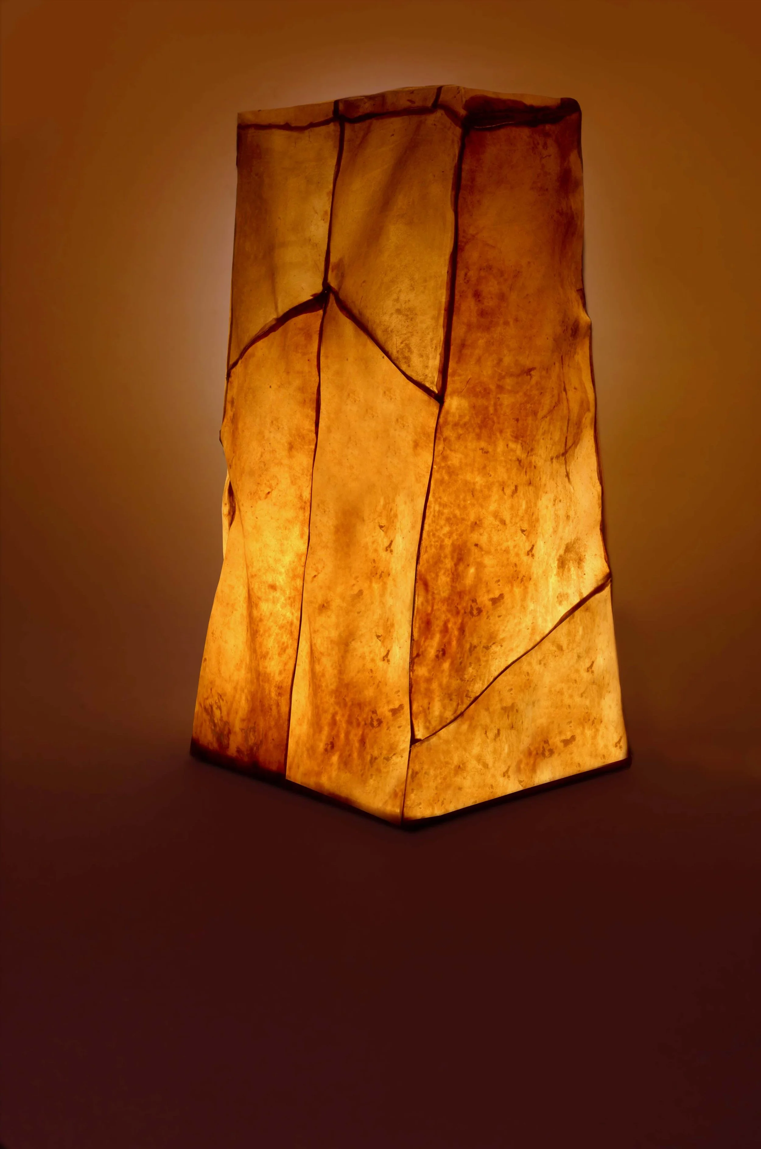 A tall, rectangular paper lantern with a warm orange glow, featuring irregularly shaped panels and visible seams, standing on a flat surface.