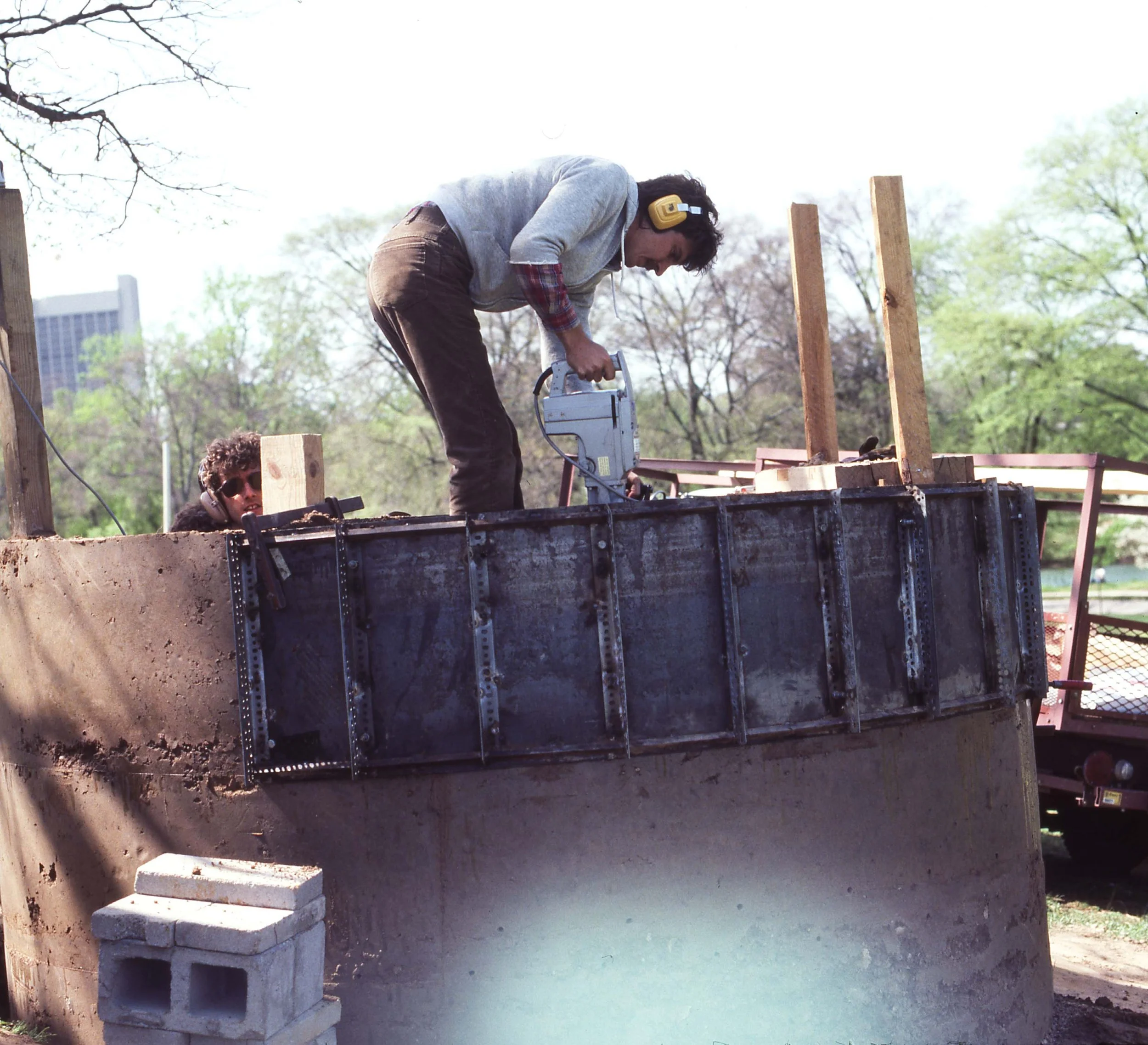 Two men working on a construction site outdoors, one using a saw on a wooden frame while the other watches, with trees and a building in the background.