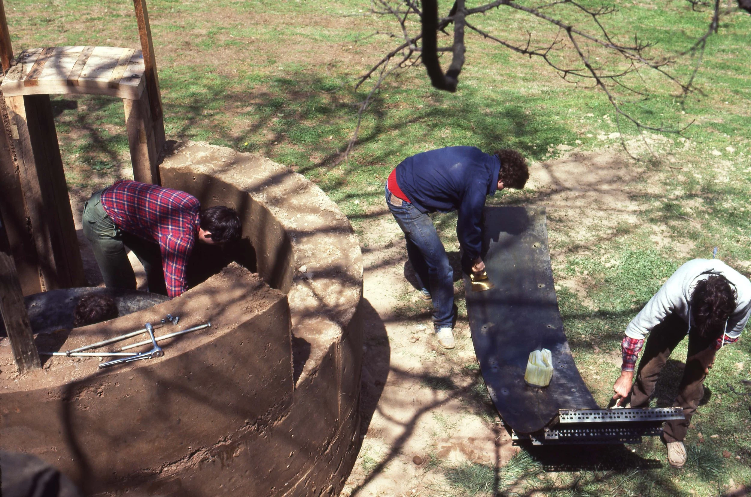 Three people working together outdoors on a construction or gardening project, involving digging and handling materials, with tools and a wheelbarrow.