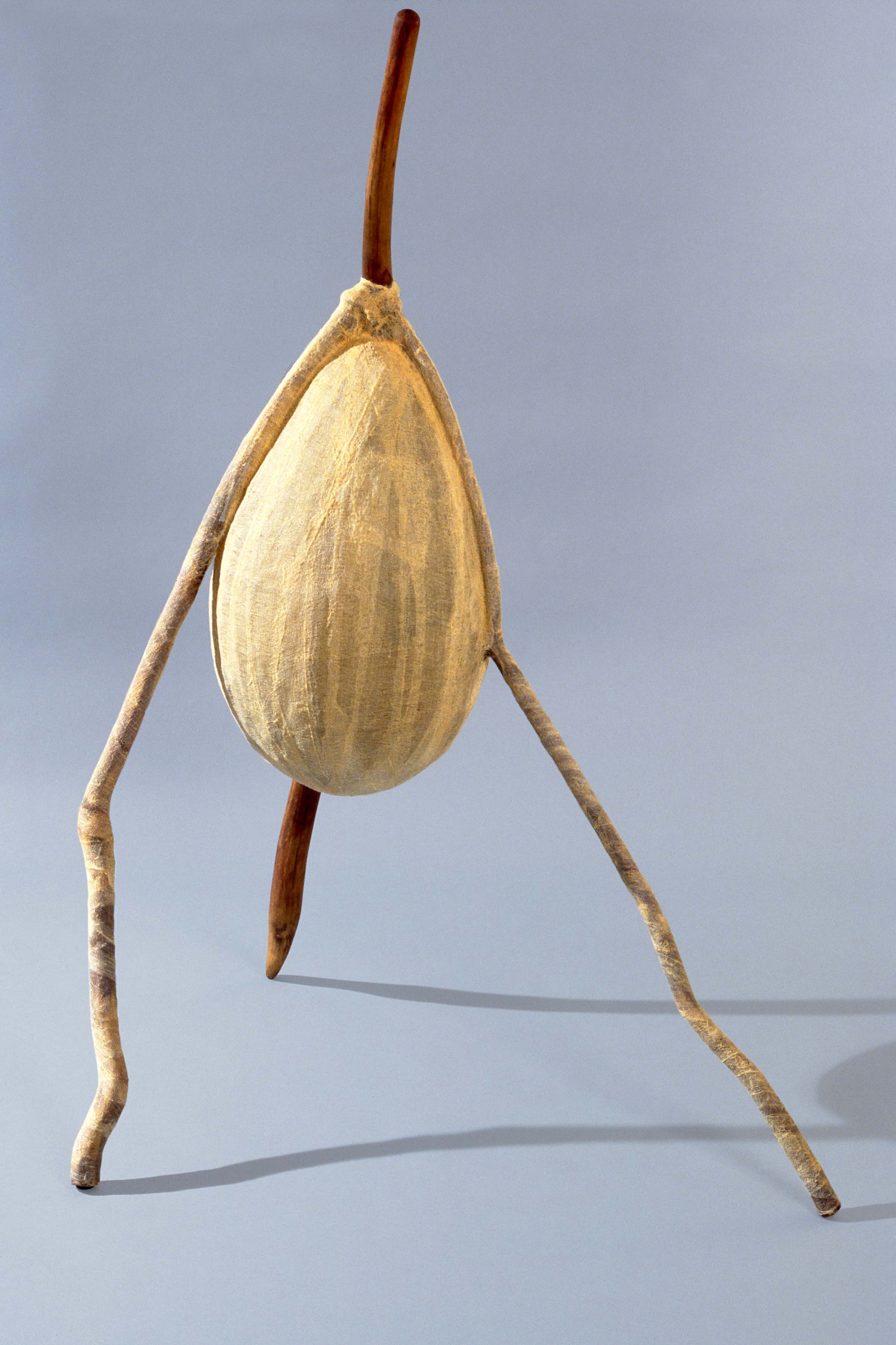 A creative sculpture resembling a walking stick figure, made from a large almond nut as the body, with dry twigs as limbs and a small twig as an arm, set against a neutral gray background.