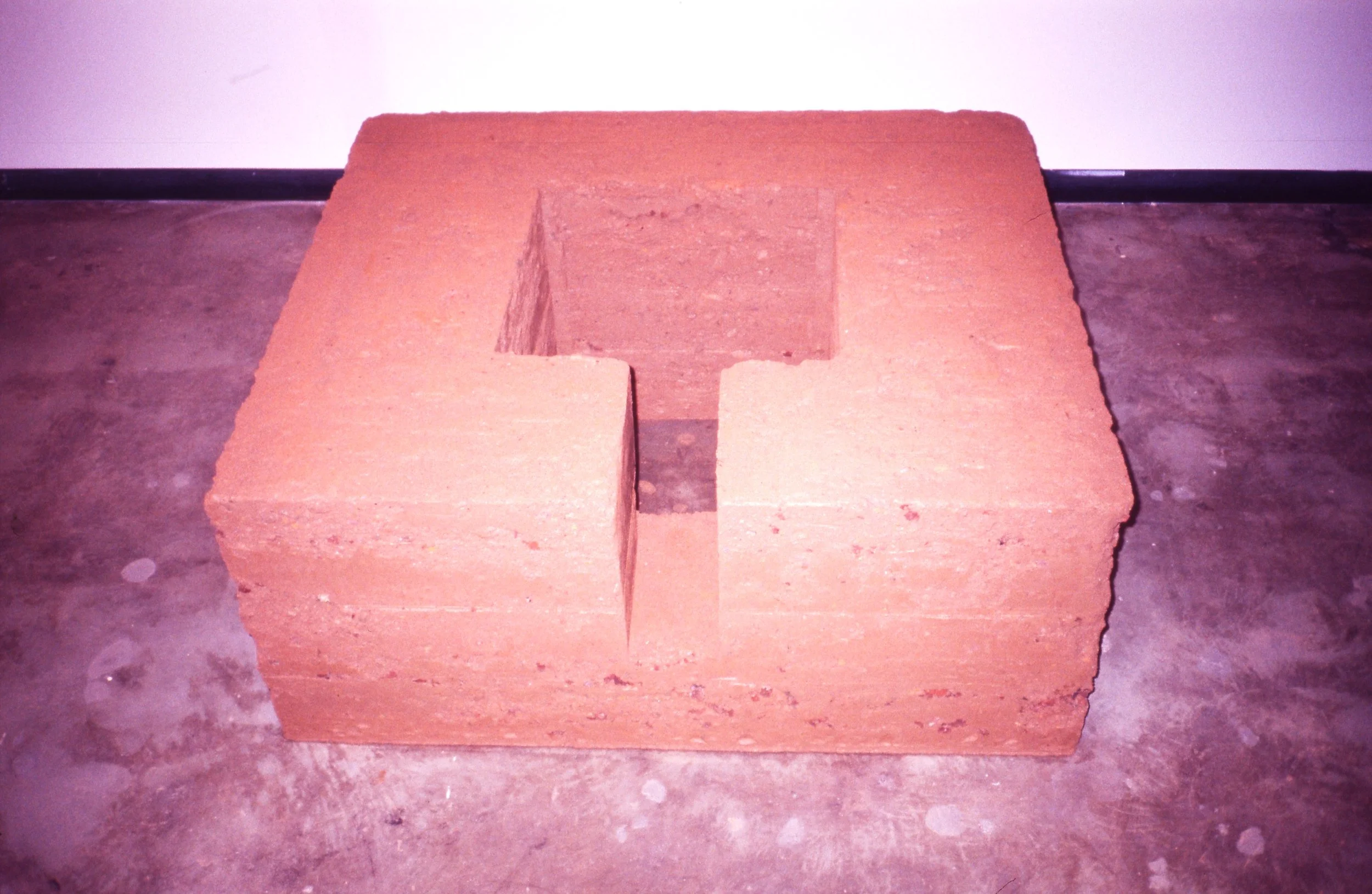 A large, hollow, rectangular brick with a cut-out section on one side, placed on a concrete floor next to a wall.
