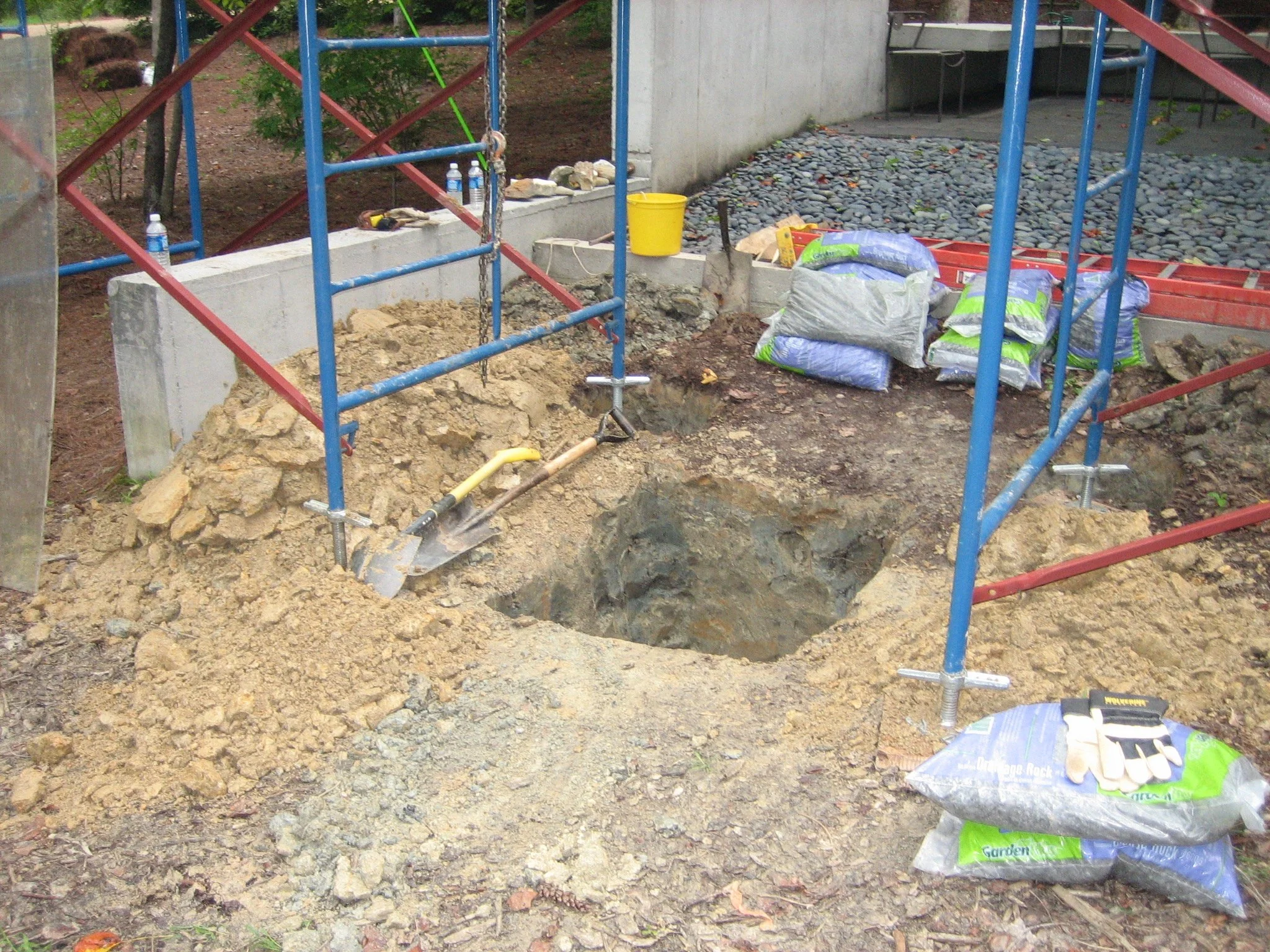A construction site with an open hole in the ground, surrounded by metal scaffolding and construction tools. There are bags of soil or gravel, a bucket, and a shovel next to the hole.