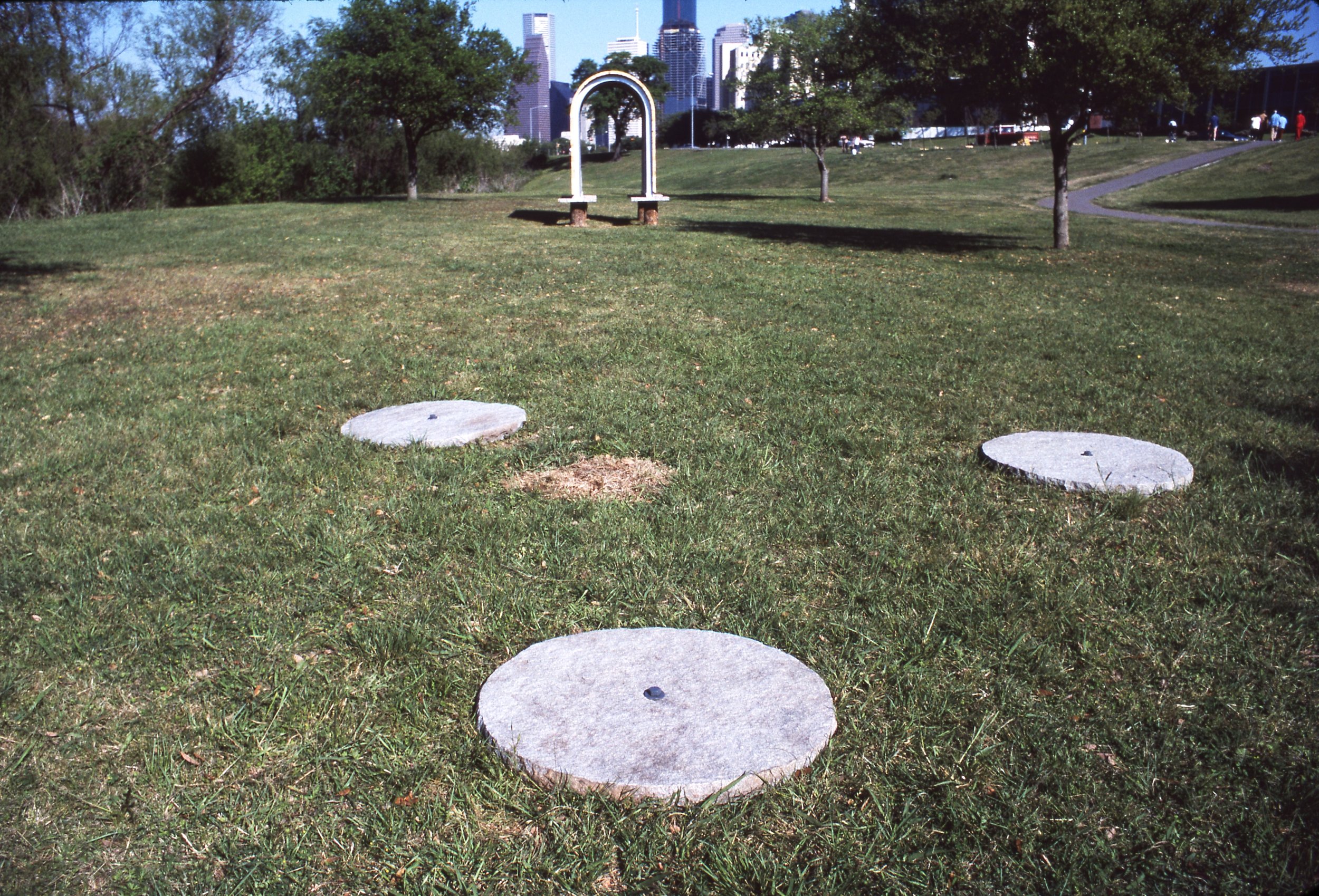 Grass field with three large round concrete slabs laid out in a line, with a park arch and city skyline in the background.