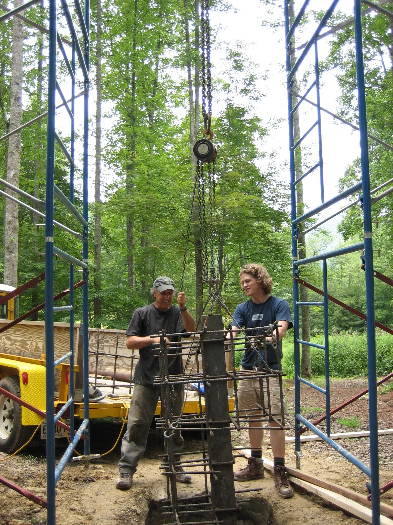 Two people working together on a construction project outdoors in a wooded area. They are handling metal rebar and equipment, with scaffolding and a yellow trailer visible in the background.