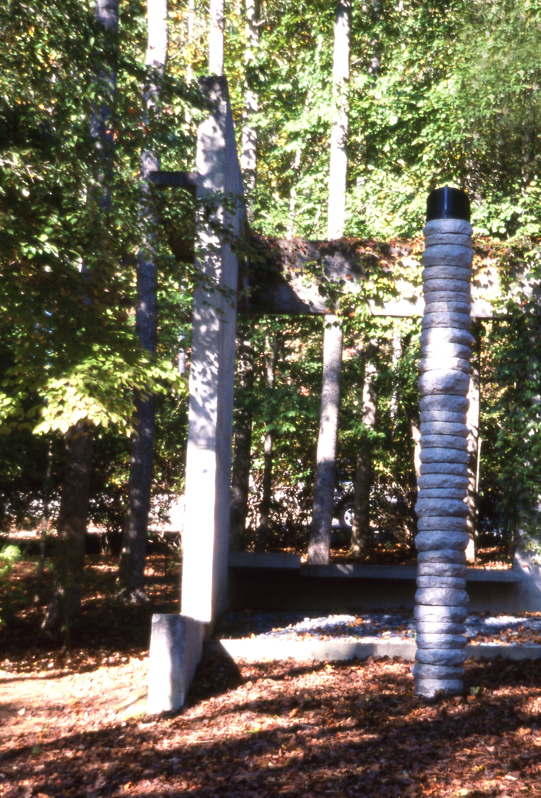 Outdoor sculpture featuring a vertical, stacked stone pillar and a cross made of light-colored stone, set amidst trees and fallen leaves.