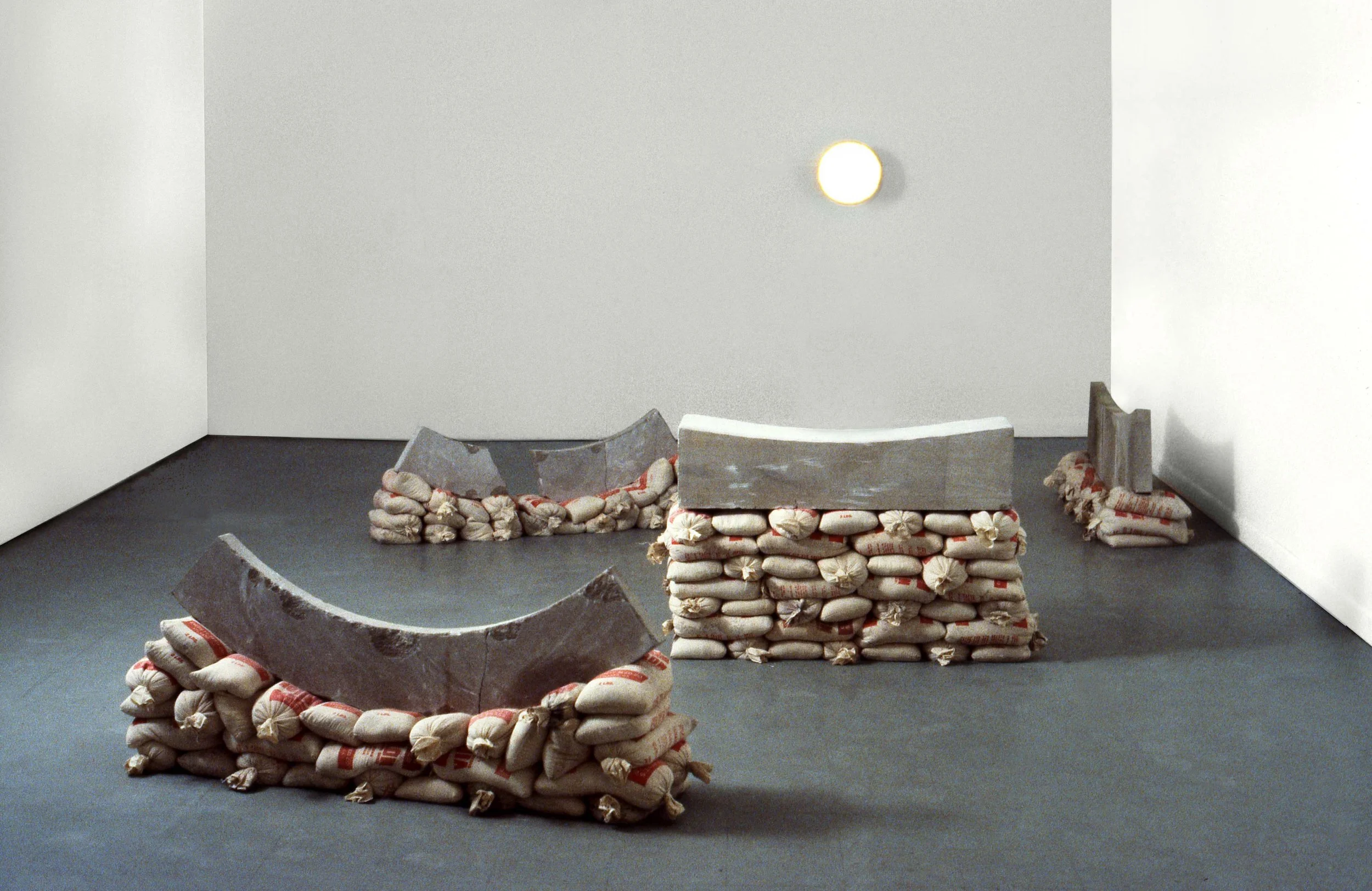 Gallery space with white walls and concrete floor, featuring sculptures made of sandbags and curved concrete pieces arranged in a geometric layout, illuminated by a single wall-mounted light.