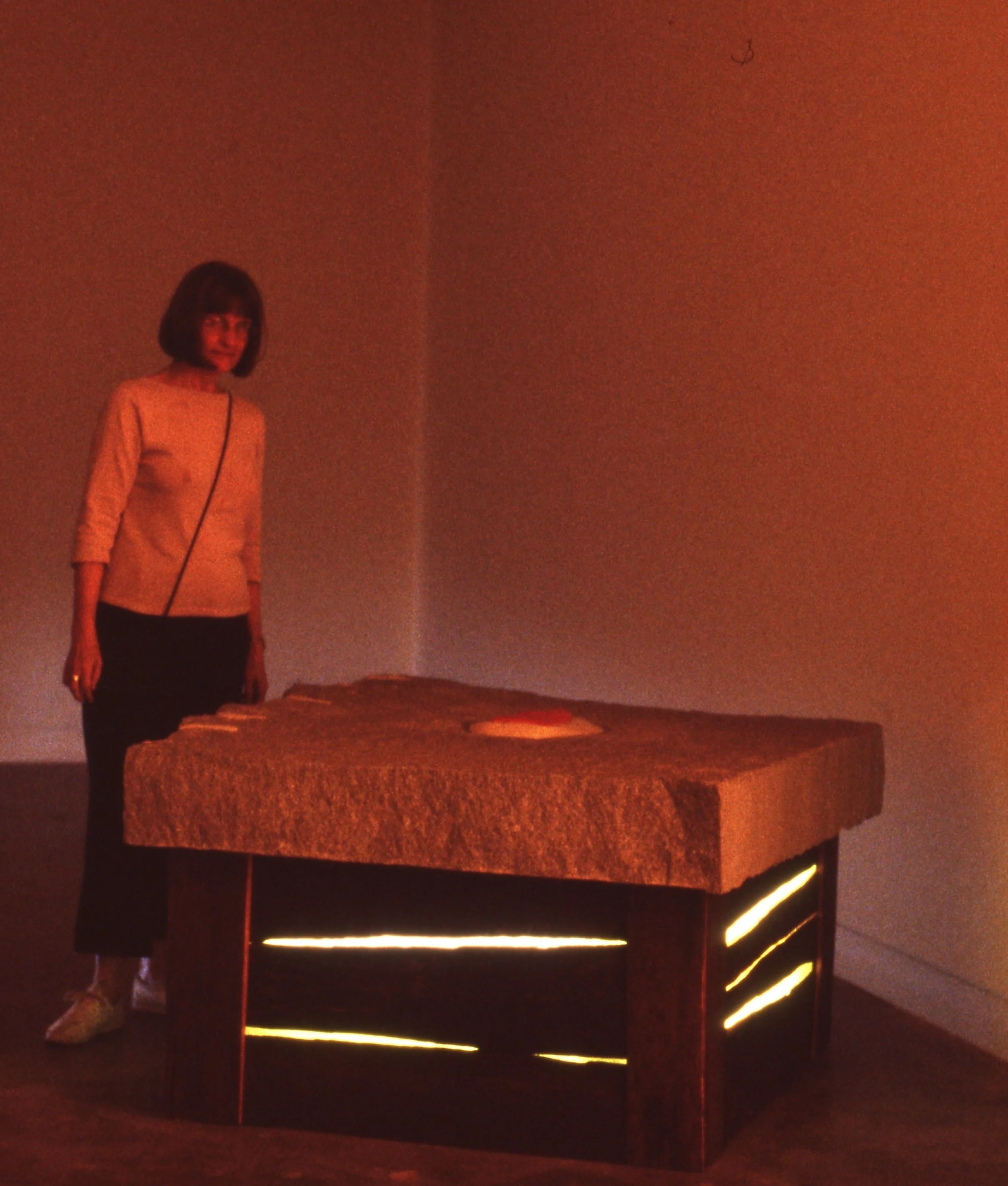 A woman standing next to a large table with a textured surface and a display of light underneath, in a dimly lit room with an orange hue.