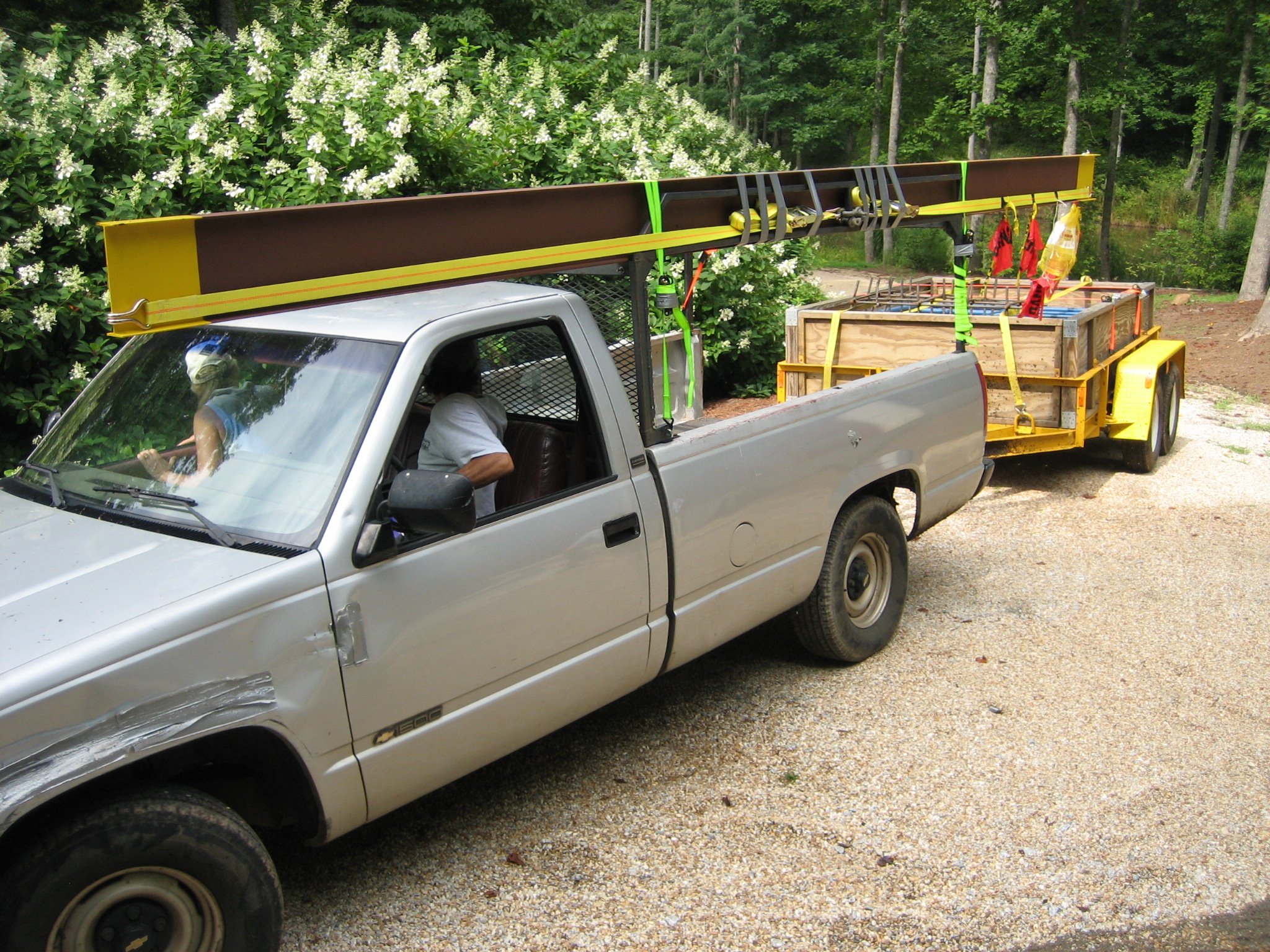 A gray pickup truck parked on a gravel driveway with a yellow utility trailer attached, carrying construction materials.