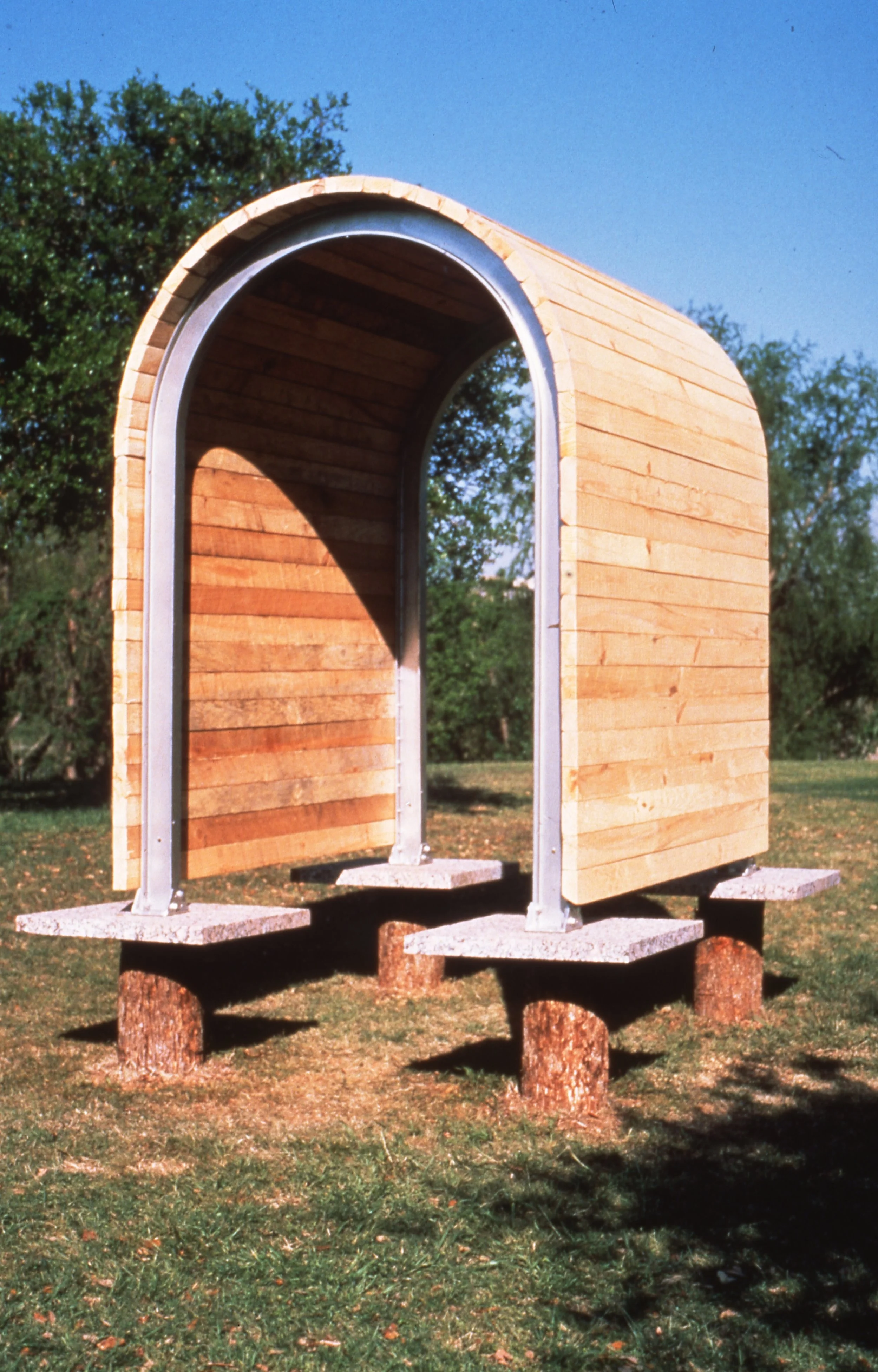 A small wooden structure with an arched top, elevated on tree stumps, outdoors on a grassy area with trees and a clear blue sky in the background.