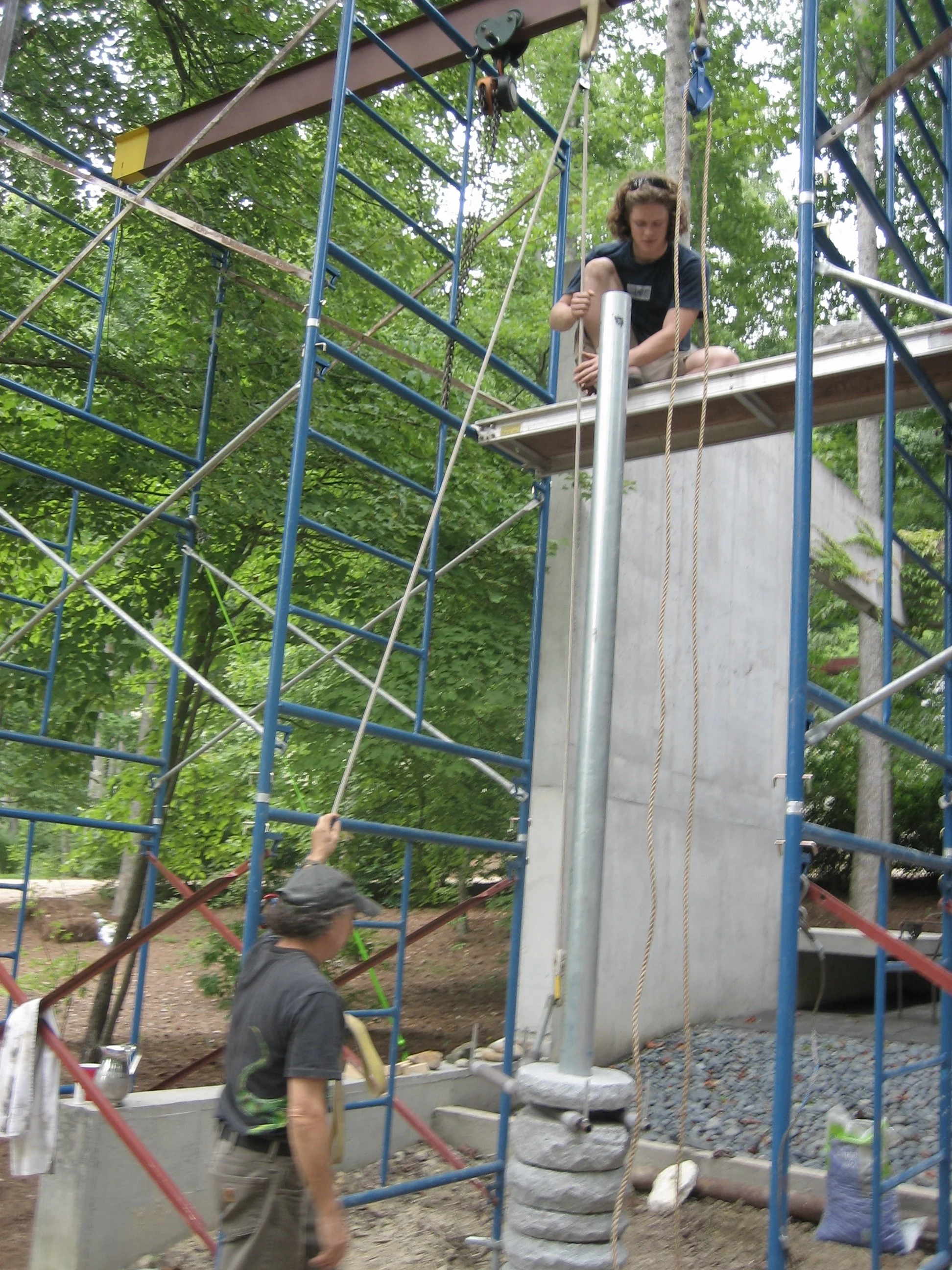 Two people on a metal climbing structure with a worker below, surrounded by trees.