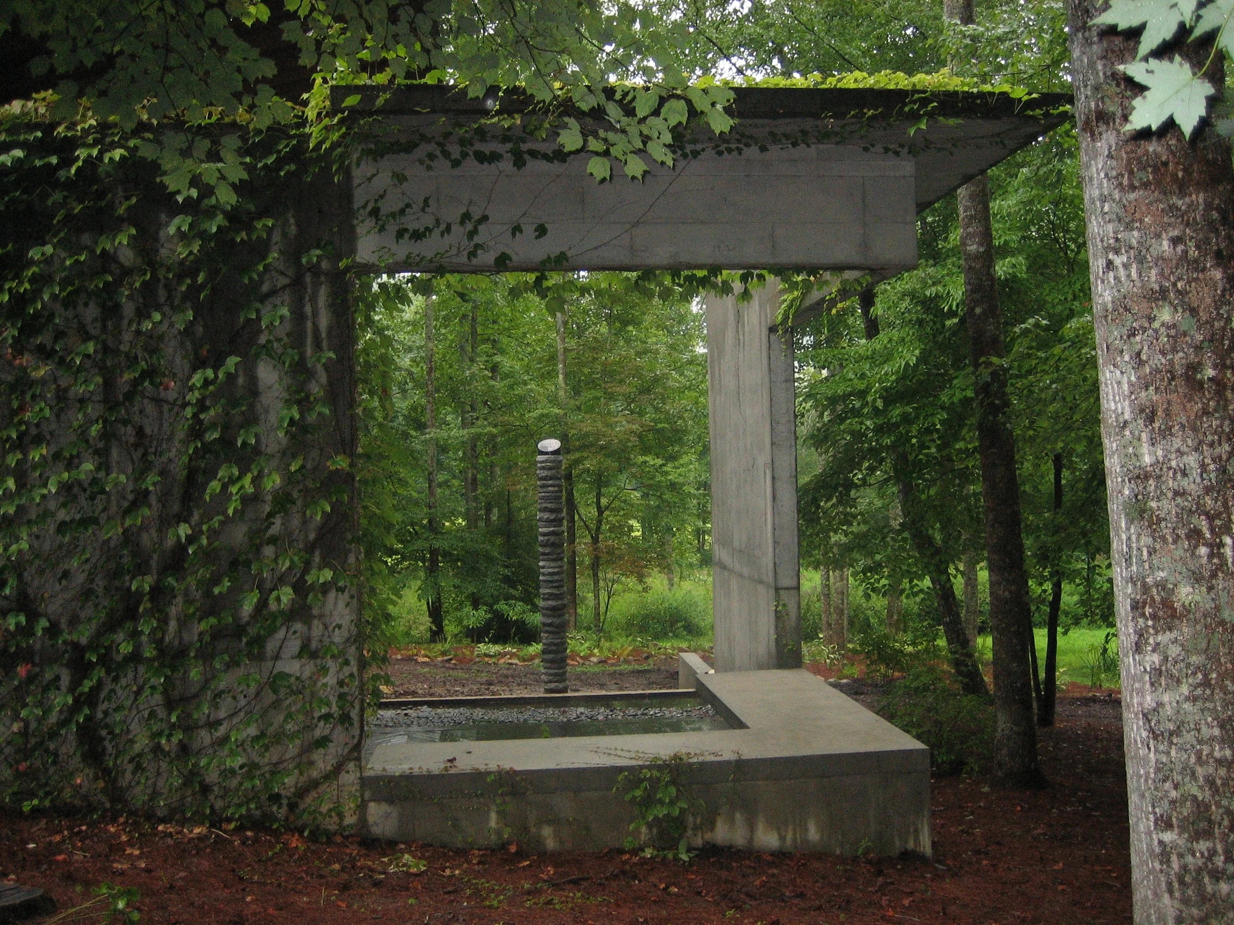 Modern concrete sculpture in a forested area with dirt ground, trees, and green foliage.