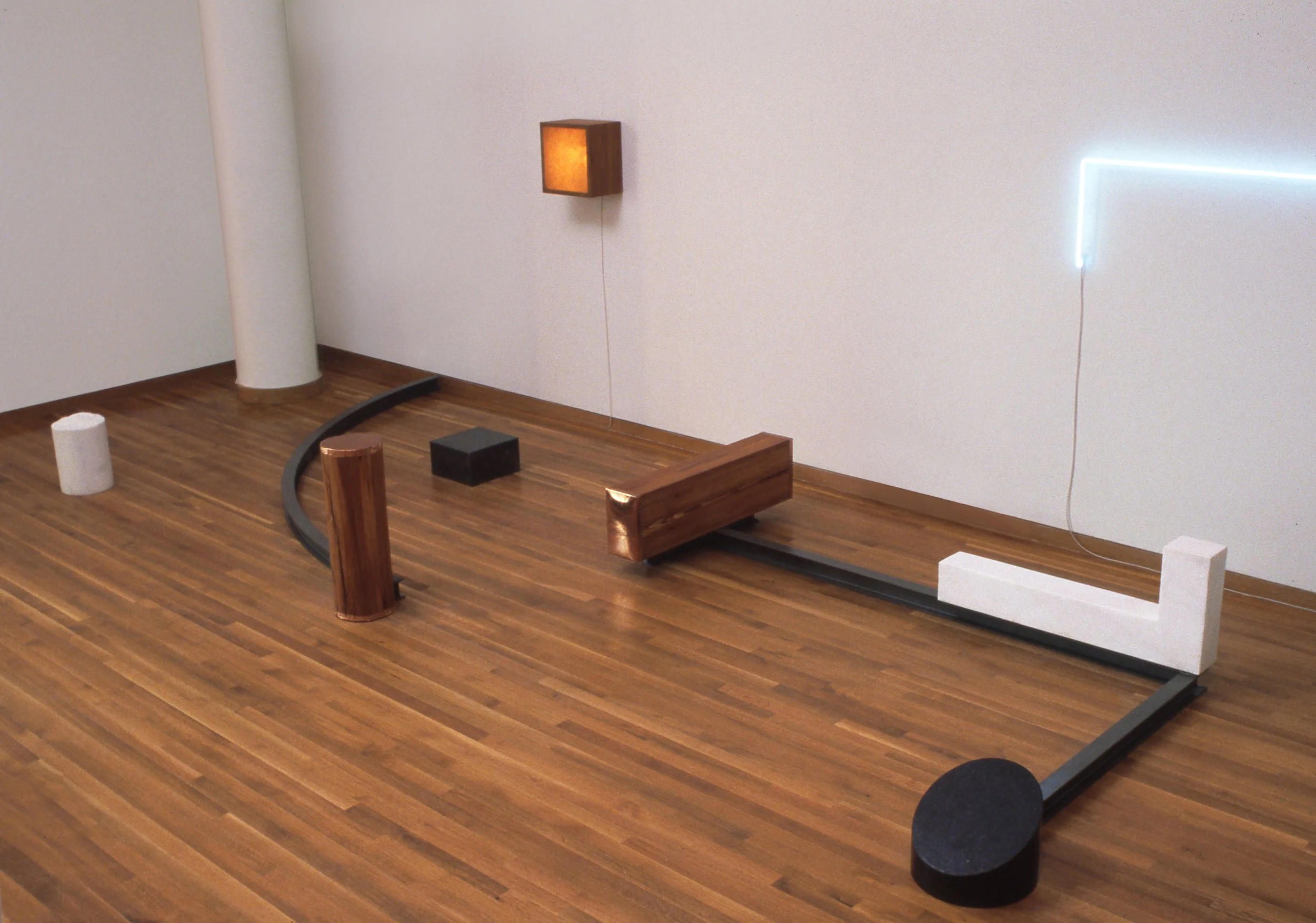 Abstract art installation with wooden and black elements on a wooden floor, featuring a curved black line, a white L-shaped block, a polished wooden rectangle, a cylindrical black piece, a vertical wood slab, a small black box, and a square wall-mounted light fixture.
