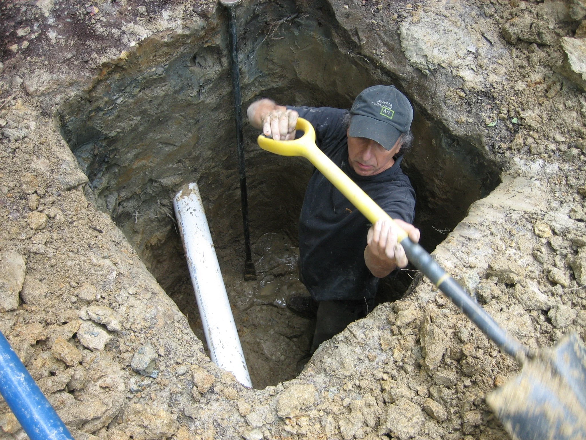 A man digging a hole with a shovel in an outdoor dirt area, wearing a cap and black shirt.