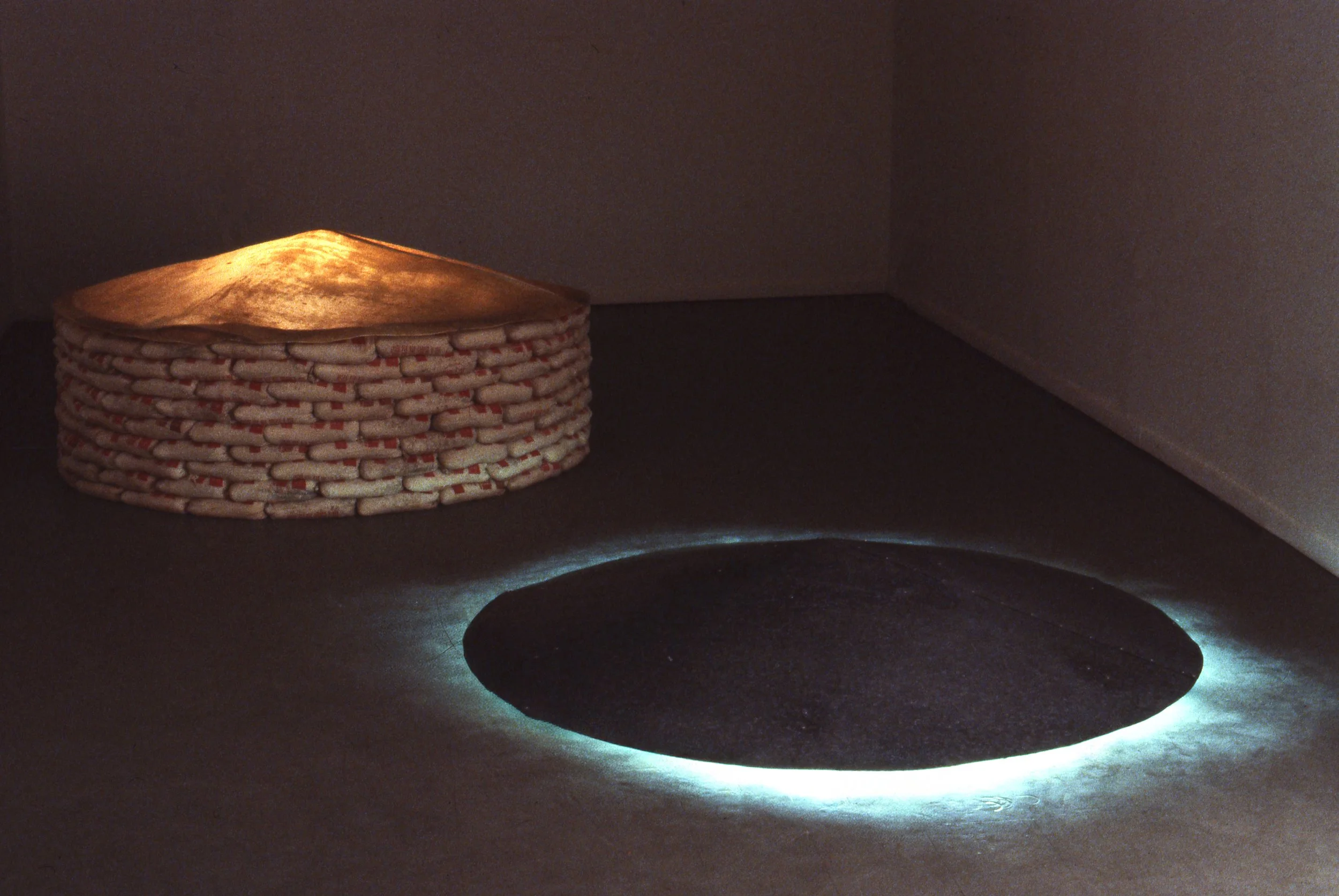 An art installation with a round, layered structure resembling a woven basket, and a circular cutout on the floor with lighting around its edge in a dark room.