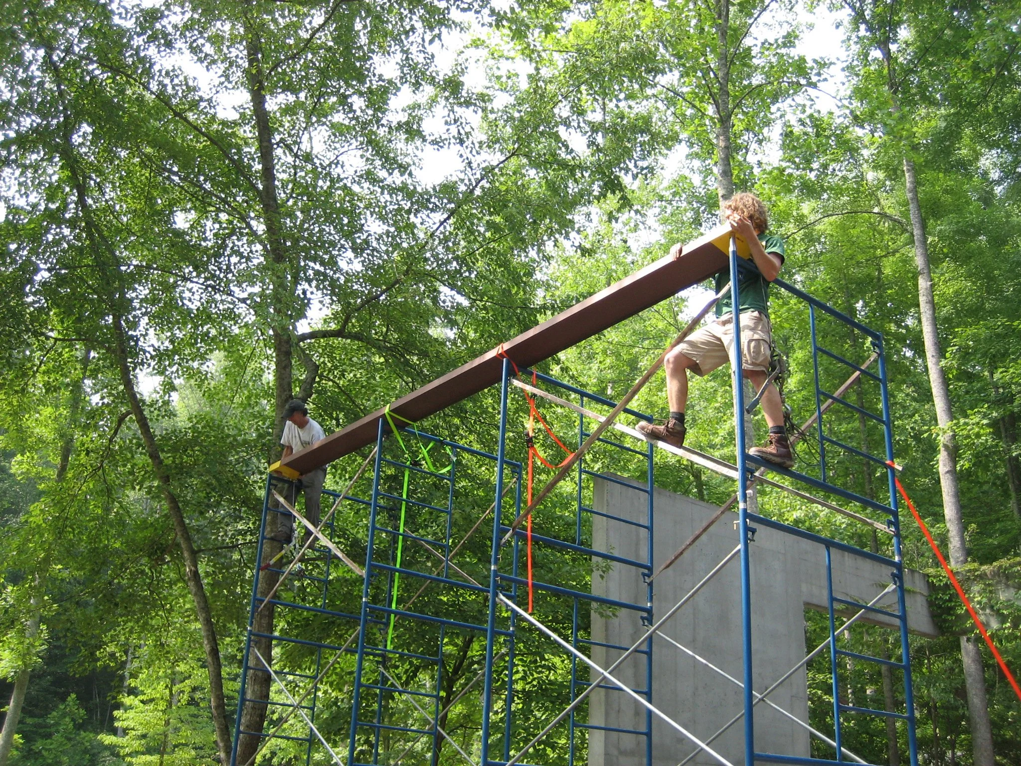 Two people are constructing a playground structure in a forested area. One person is on the left, working on the scaffolding, while the other is on the right, holding a long wooden beam on top of the structure.