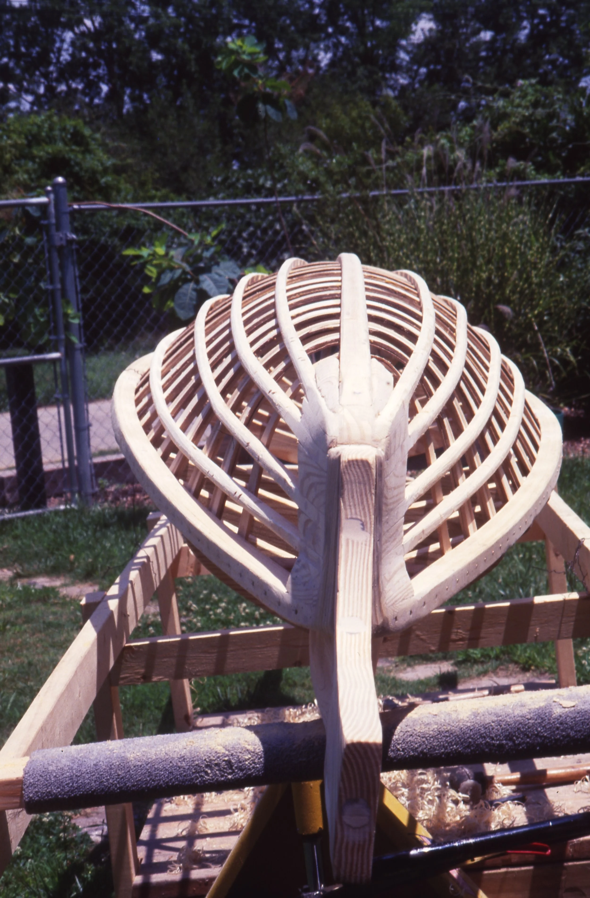 Wooden boat skeleton frame for a small boat or kayak under construction outdoors with garden and fence in background.